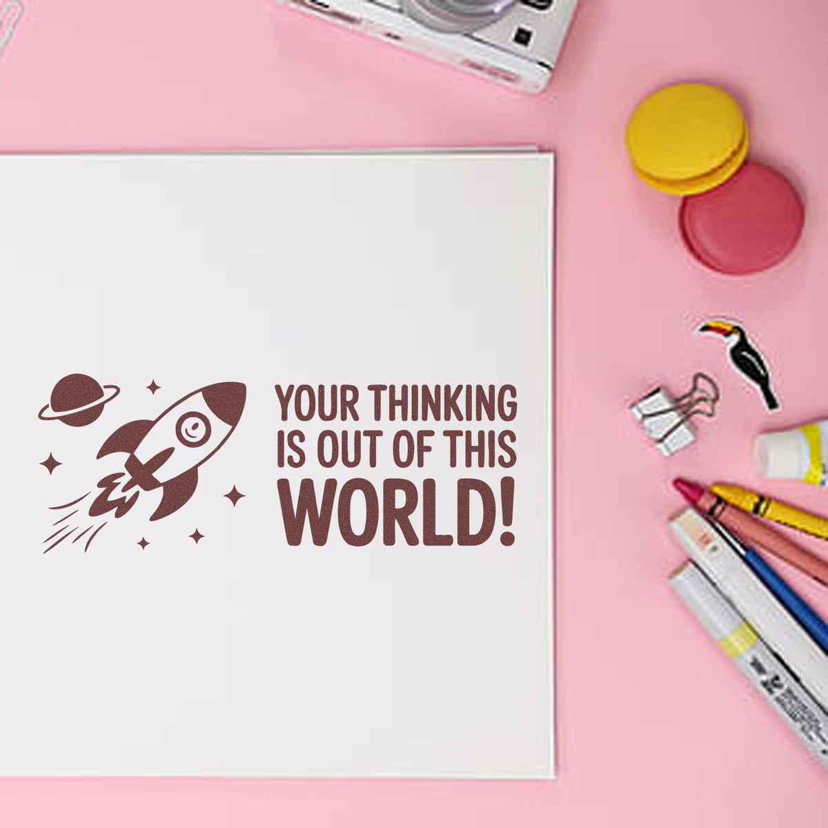 Space rocket rubber stamp reading Your thinking is out of this world! on a notebook with craft supplies on a pink background.