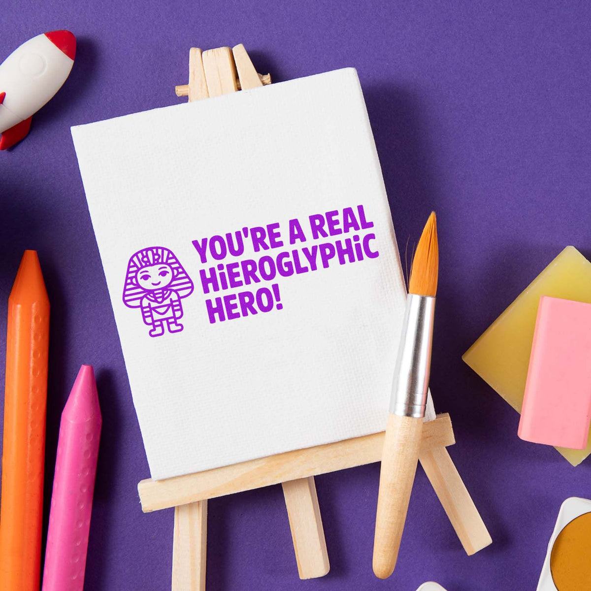 Custom rubber stamp impression reading Youre a real hieroglyphic hero! on canvas easel with art supplies.