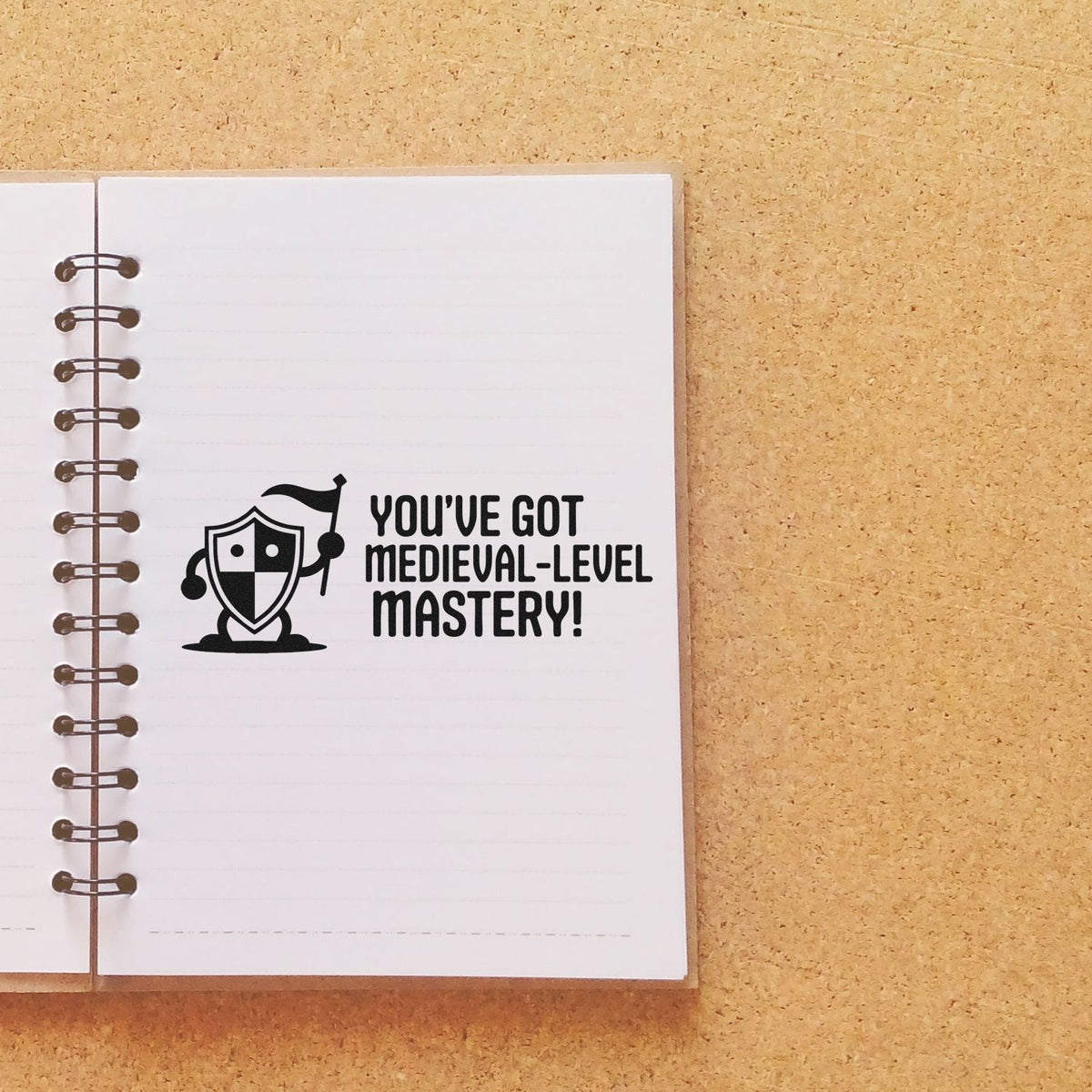 Medieval-themed rubber stamp reading Youve Got Medieval-Level Mastery! with knight shield on notebook page.