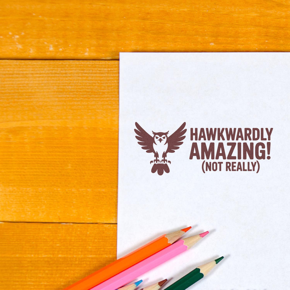 Custom owl rubber stamp reading Hawkwardly Amazing (Not Really) on white paper with colored pencils on a wooden desk.