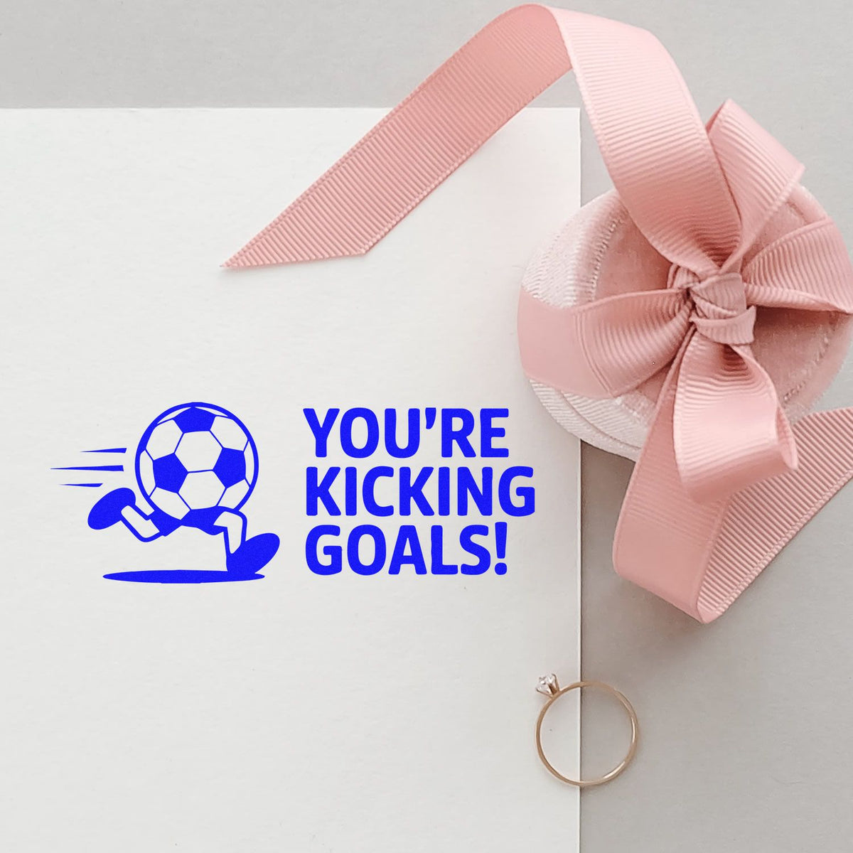 Soccer rubber stamp reading Youre Kicking Goals! on white paper, styled with pink ribbon bow and gift packaging.