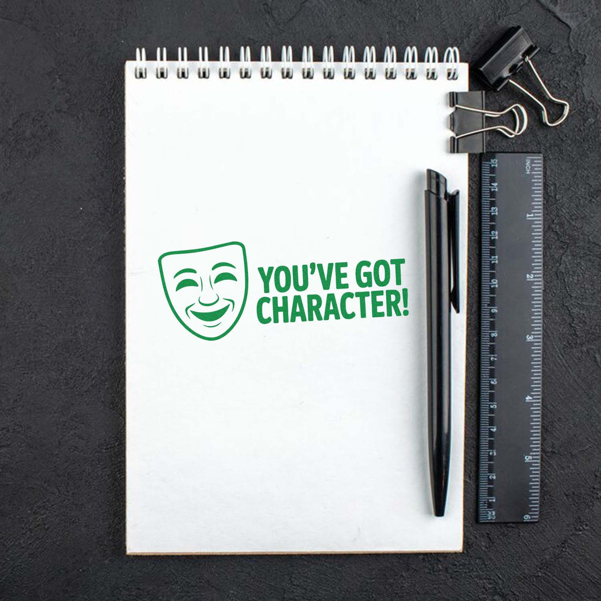Custom rubber stamp mockup with green comedy mask logo and Youve Got Character! on a spiral notebook.