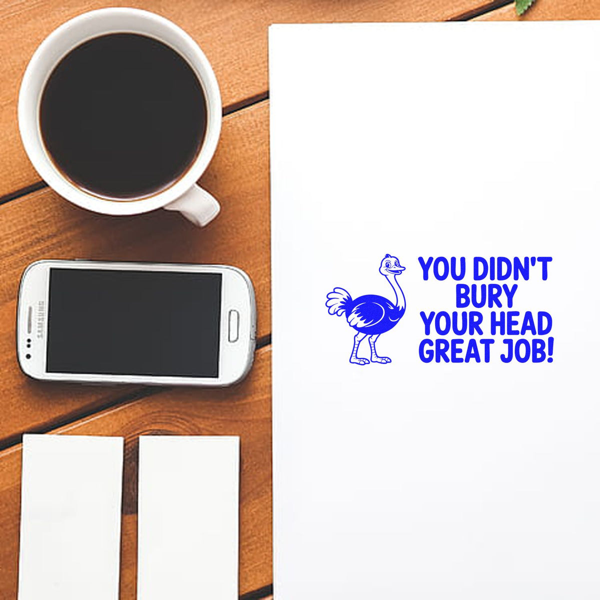 Rubber stamp mockup with ostrich graphic and You Didnt Bury Your Head Great Job! message on paper beside coffee and phone.