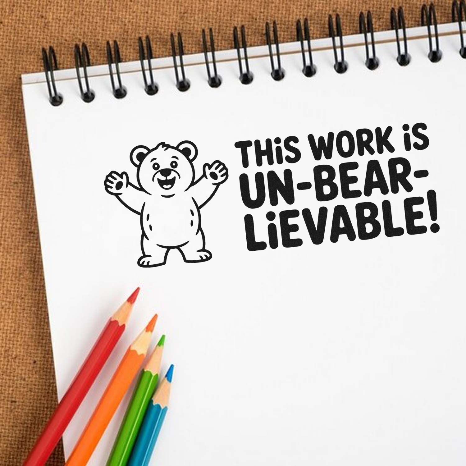 Playful bear custom rubber stamp reading This work is un-bear-lievable! on a notebook with colored pencils.