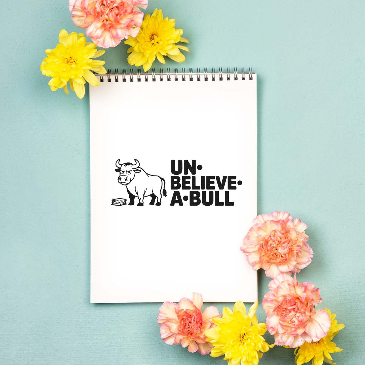 Bull pun rubber stamp design reading Un-Believe-A-Bull on a notebook, styled with flowers.