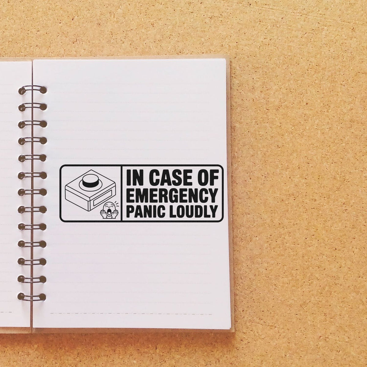 Novelty rubber stamp reading In Case of Emergency, Panic Loudly displayed on a spiral notebook page.