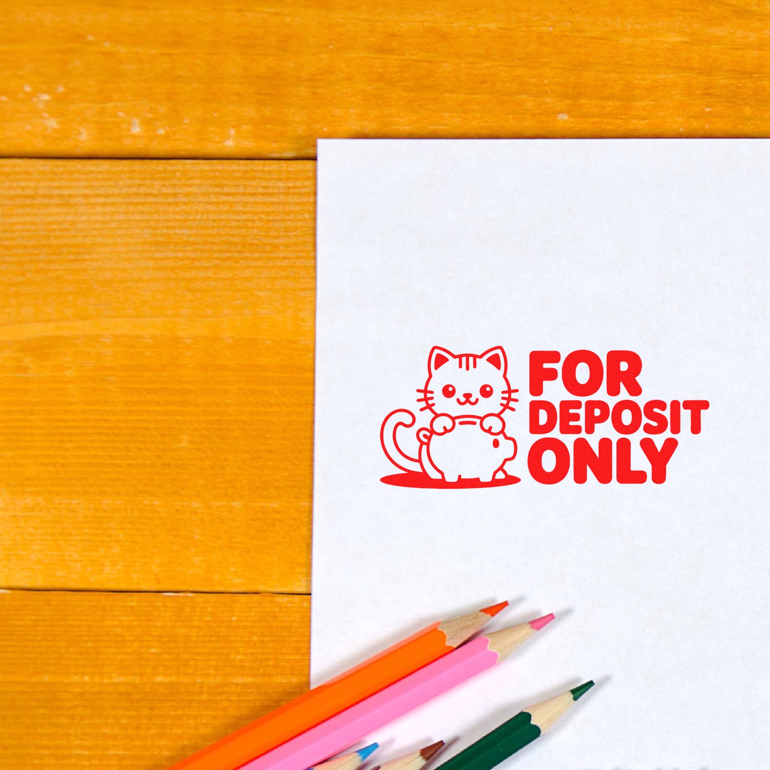Red For Deposit Only rubber stamp with cat and piggy bank icon on white paper, beside colored pencils on a wood table.