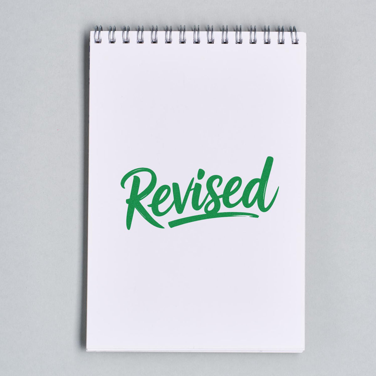 Green Revised rubber stamp impression on a spiral notebook page for office document review and approval.