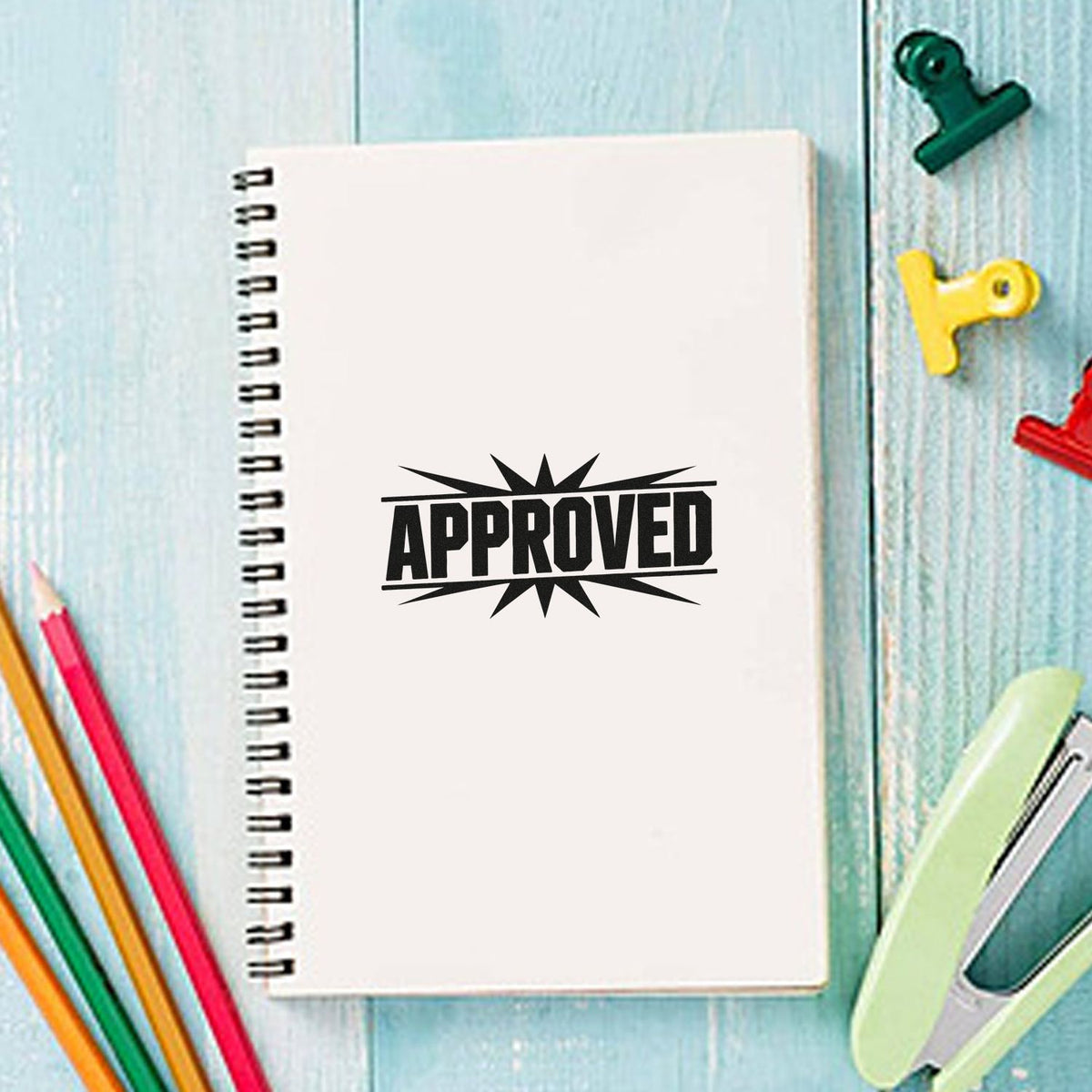 Approved rubber stamp impression on a notebook surrounded by colorful stationery and office supplies.