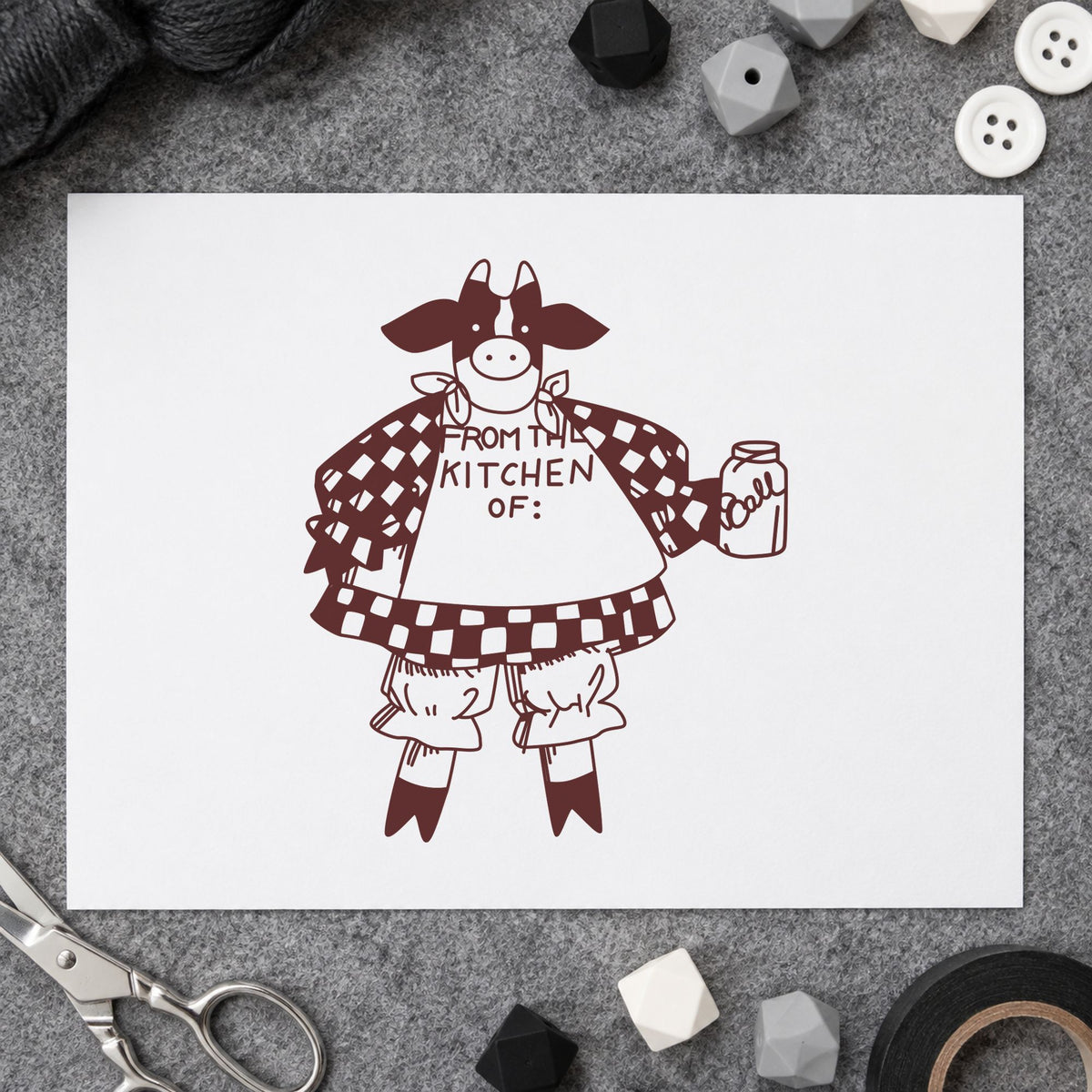 From the Kitchen Of Cow Rubber Stamp impression in brown ink on white paper in a styled craft setting.