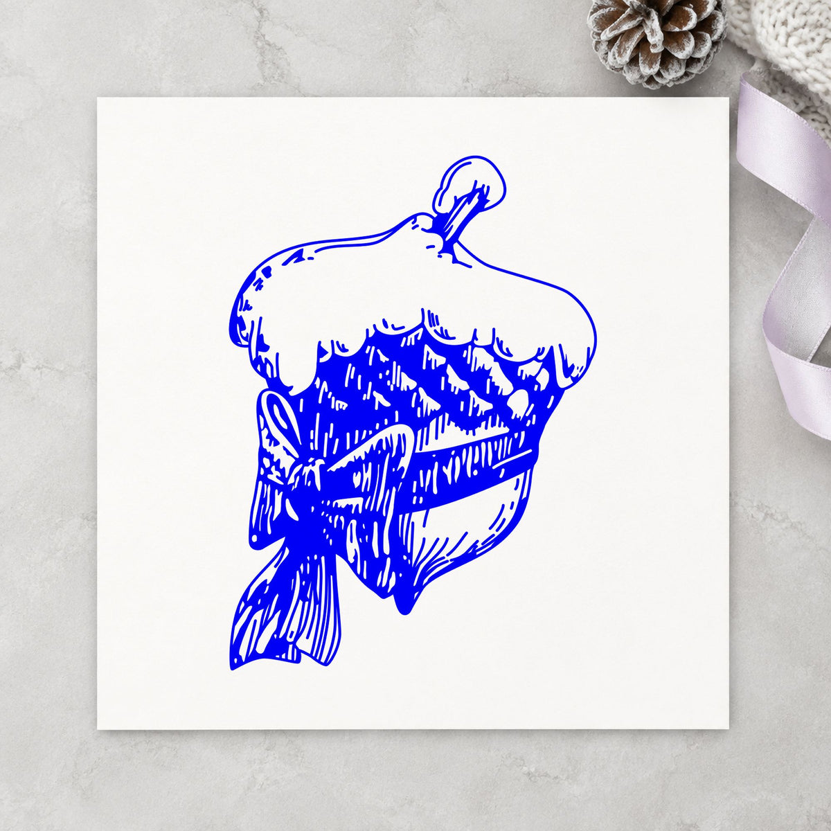 Blue winter craft stamp impression featuring the snowy acorn with bow christmas design on white paper.