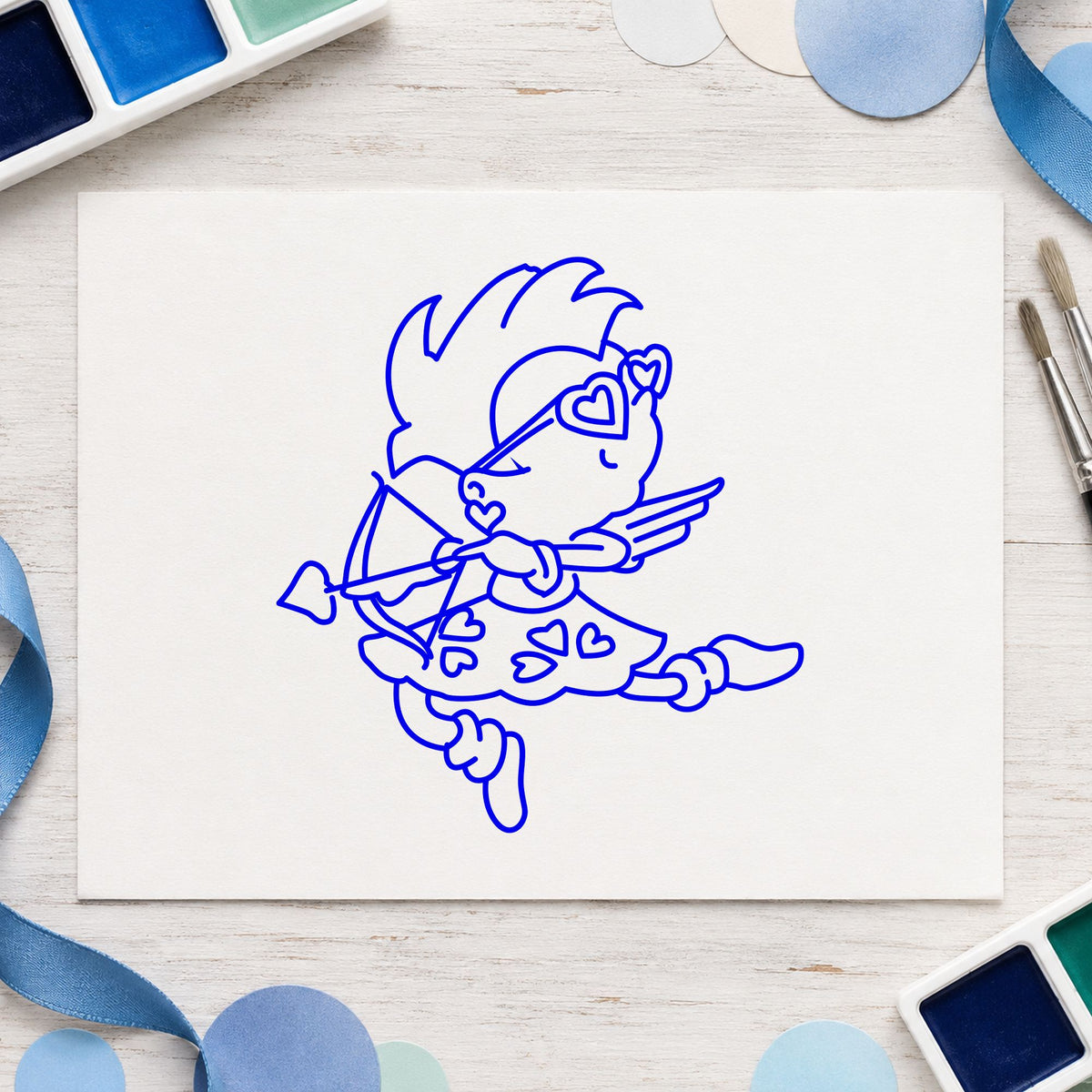 Cupid with Heart Sunglasses Rubber Stamp impression in blue ink on white paper in a styled craft setting.