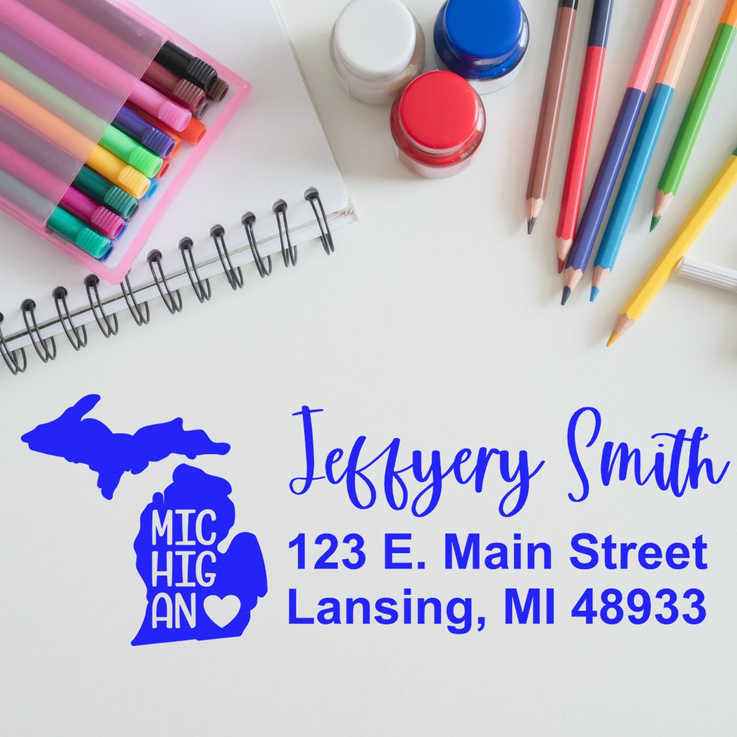 Custom return address rubber stamp with Michigan design, name and address, shown with stationery and pens for mailing use.