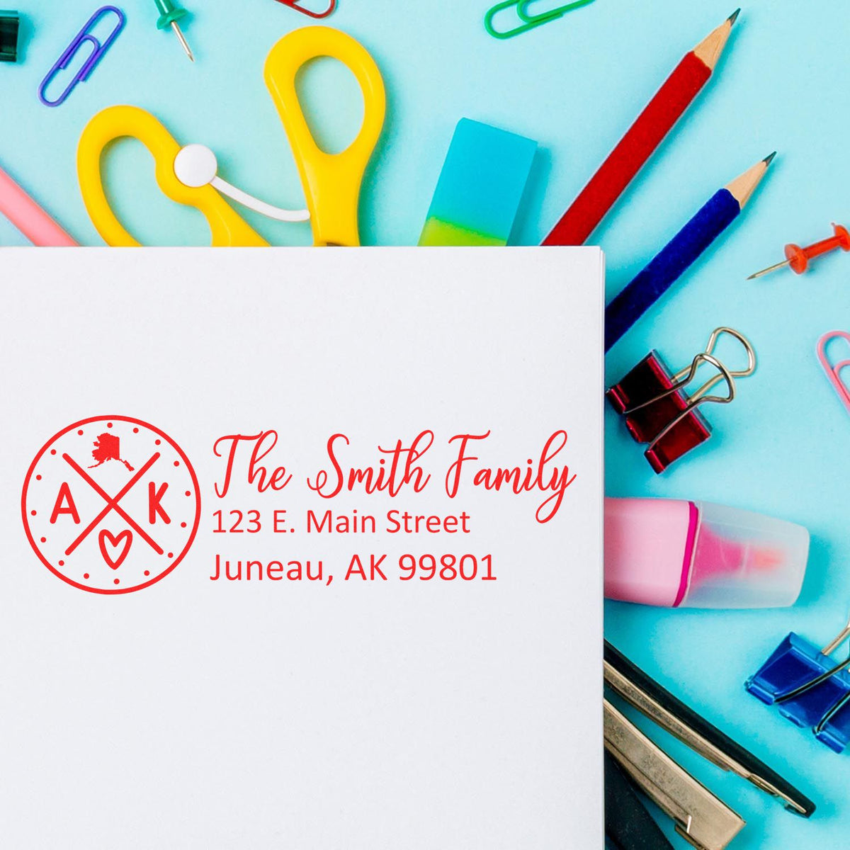 Custom family return address rubber stamp with monogram design, shown on paper with school supplies, Juneau, AK 99801.