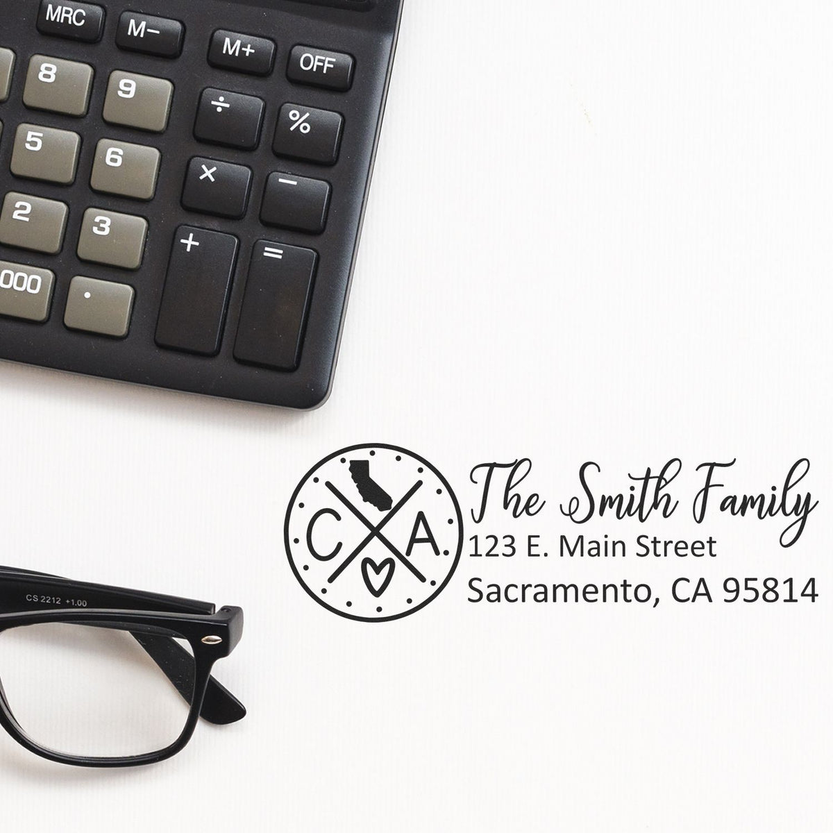 Personalized return address rubber stamp with family name and California address beside calculator and glasses on a desk