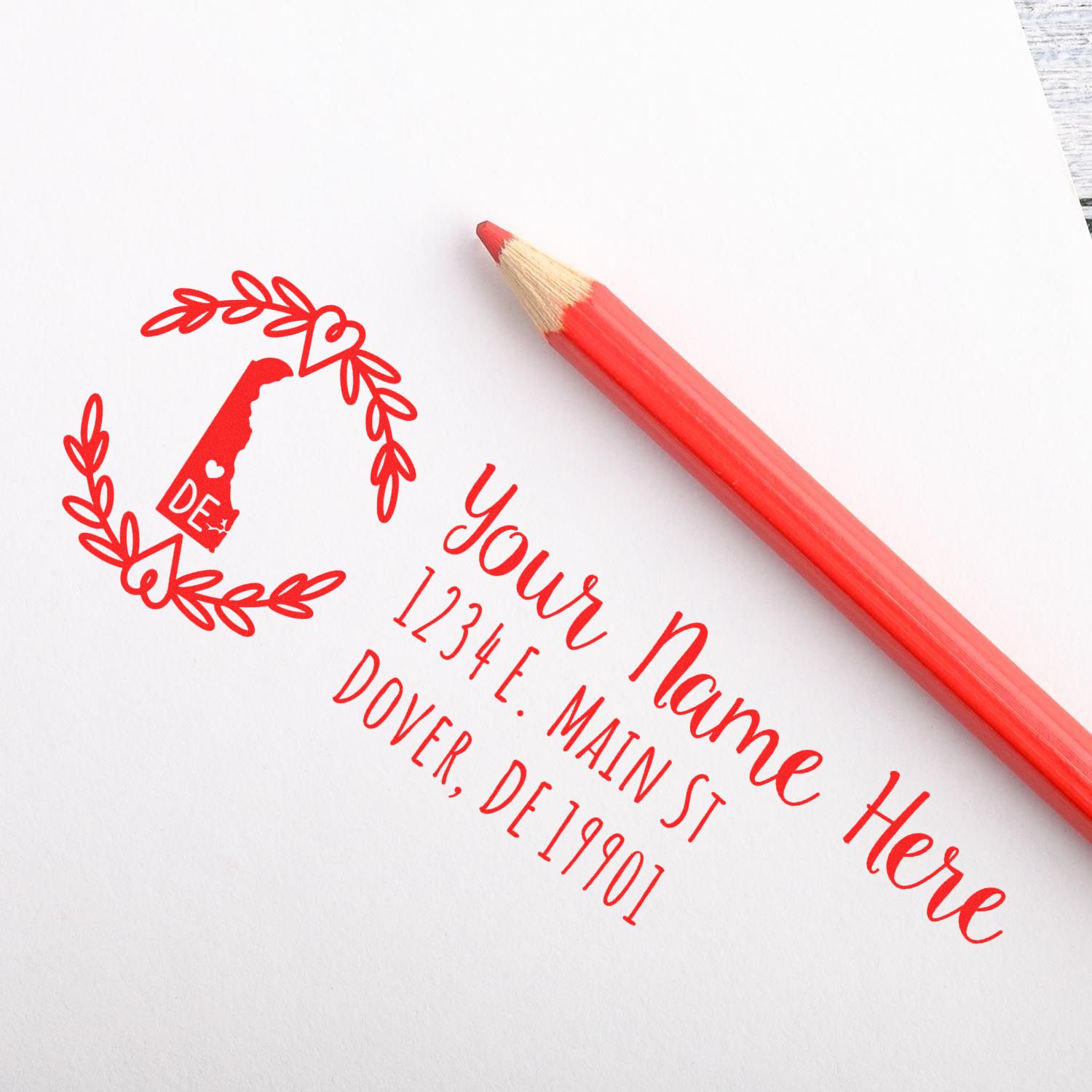 Custom return address rubber stamp with laurel wreath design, stamped in red on white paper beside a red pencil.