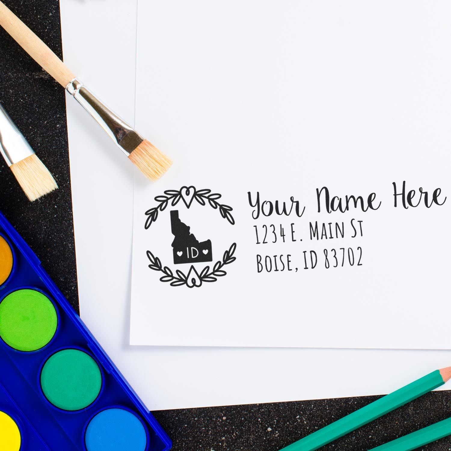 Custom Idaho return address rubber stamp with name and Boise, ID address, styled with paintbrushes and watercolor supplies.