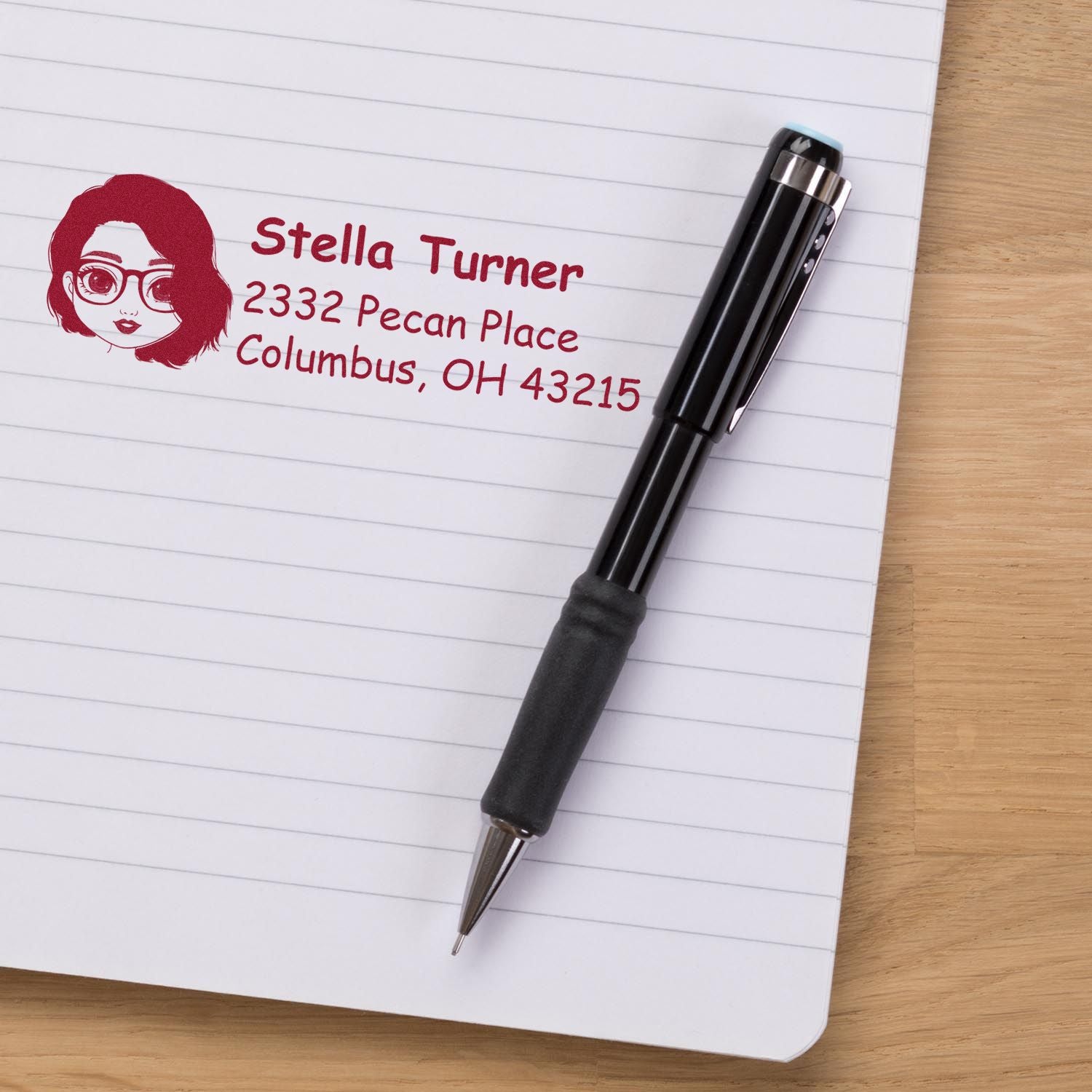 Personalized return address rubber stamp with name and address imprint on notebook beside a black pen.