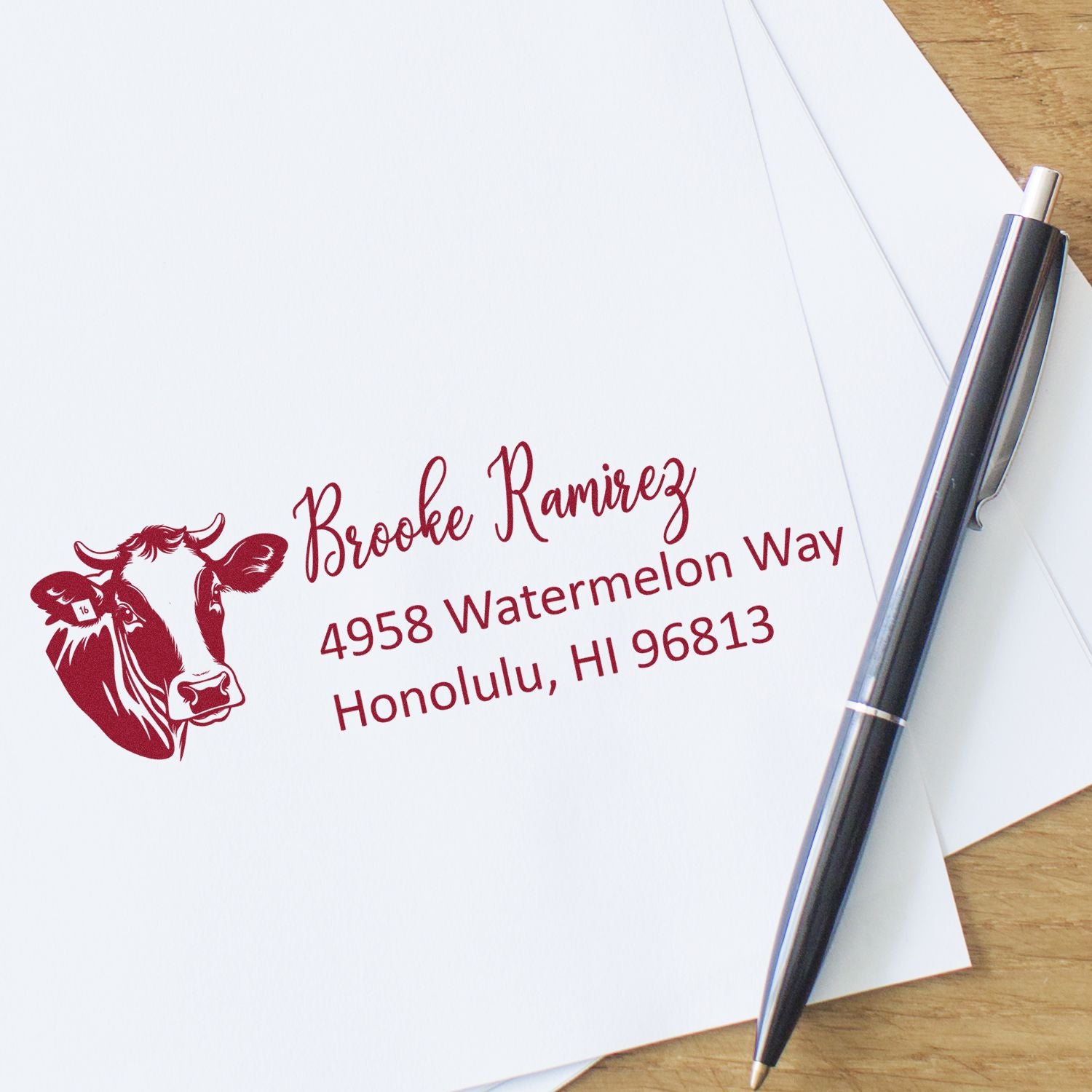 Personalized rubber return address stamp with cow illustration, name, and Honolulu address on envelope beside pen.