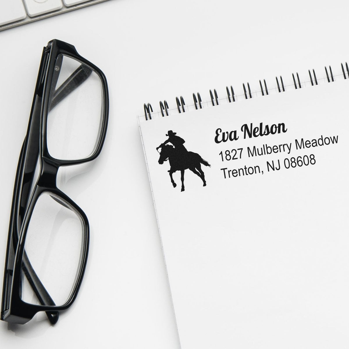 Custom return address rubber stamp with cowboy logo on spiral notebook beside eyeglasses.