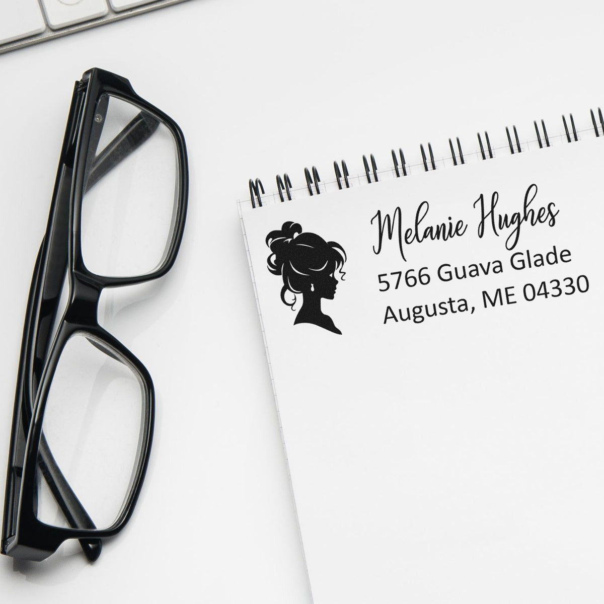 Custom return address rubber stamp with name, address and silhouette logo beside eyeglasses on a notebook.