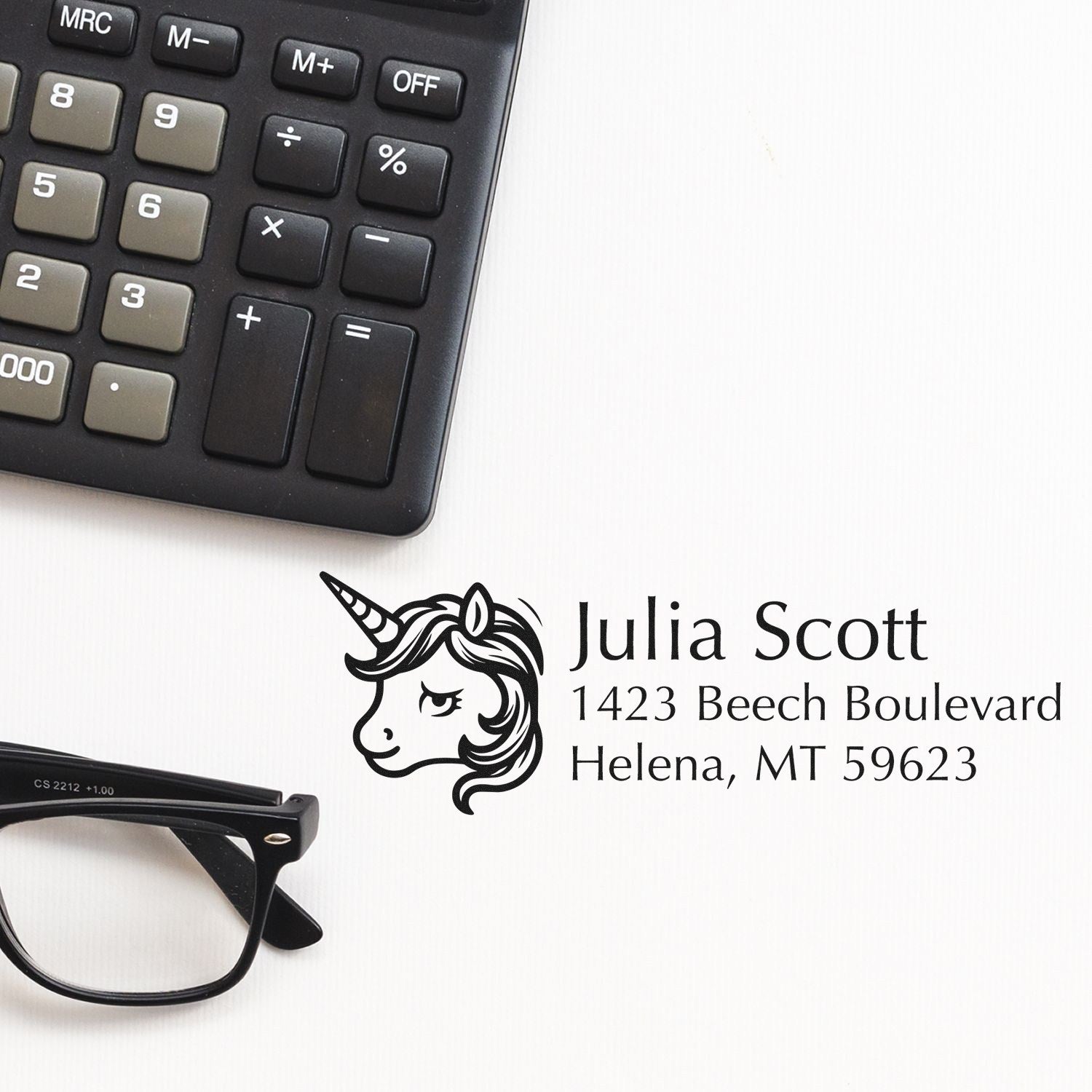 Custom unicorn address rubber stamp with name and address, displayed beside calculator and eyeglasses.