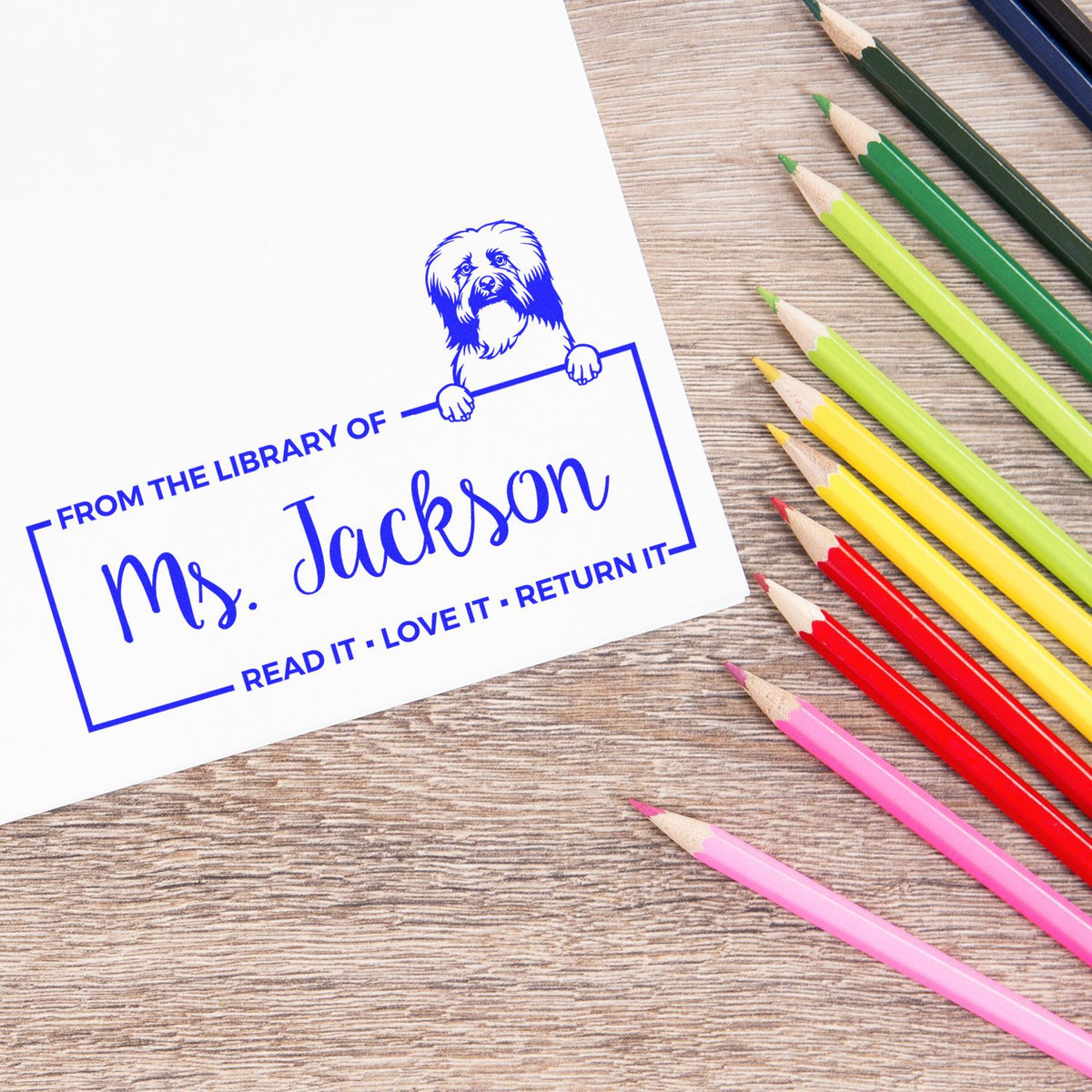 Personalized library rubber stamp impression with dog design reading From the Library of Ms. Jackson beside colored pencils.