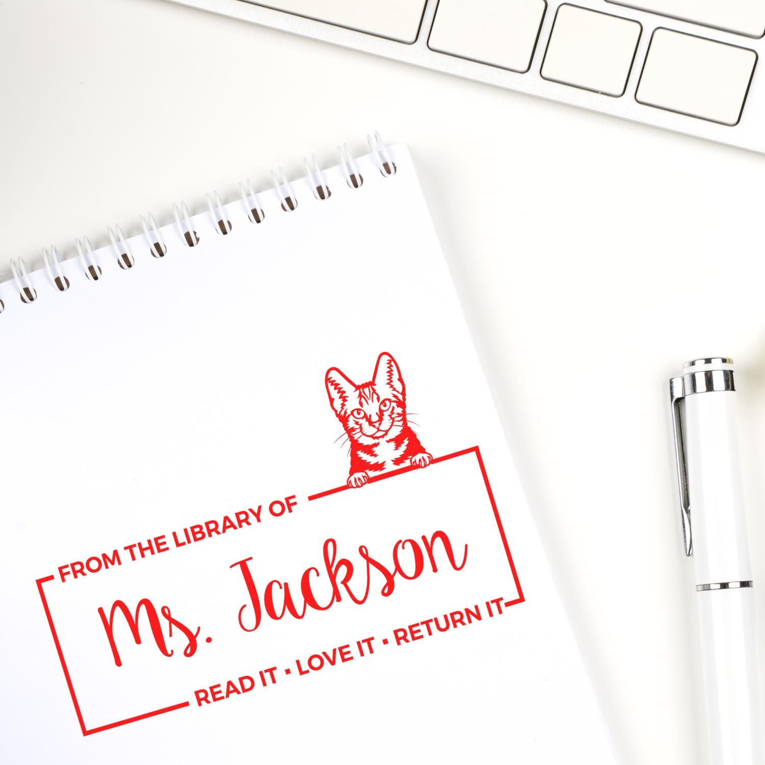 Custom rubber book stamp with cat design reading From the Library of Ms. Jackson in red on a notebook.