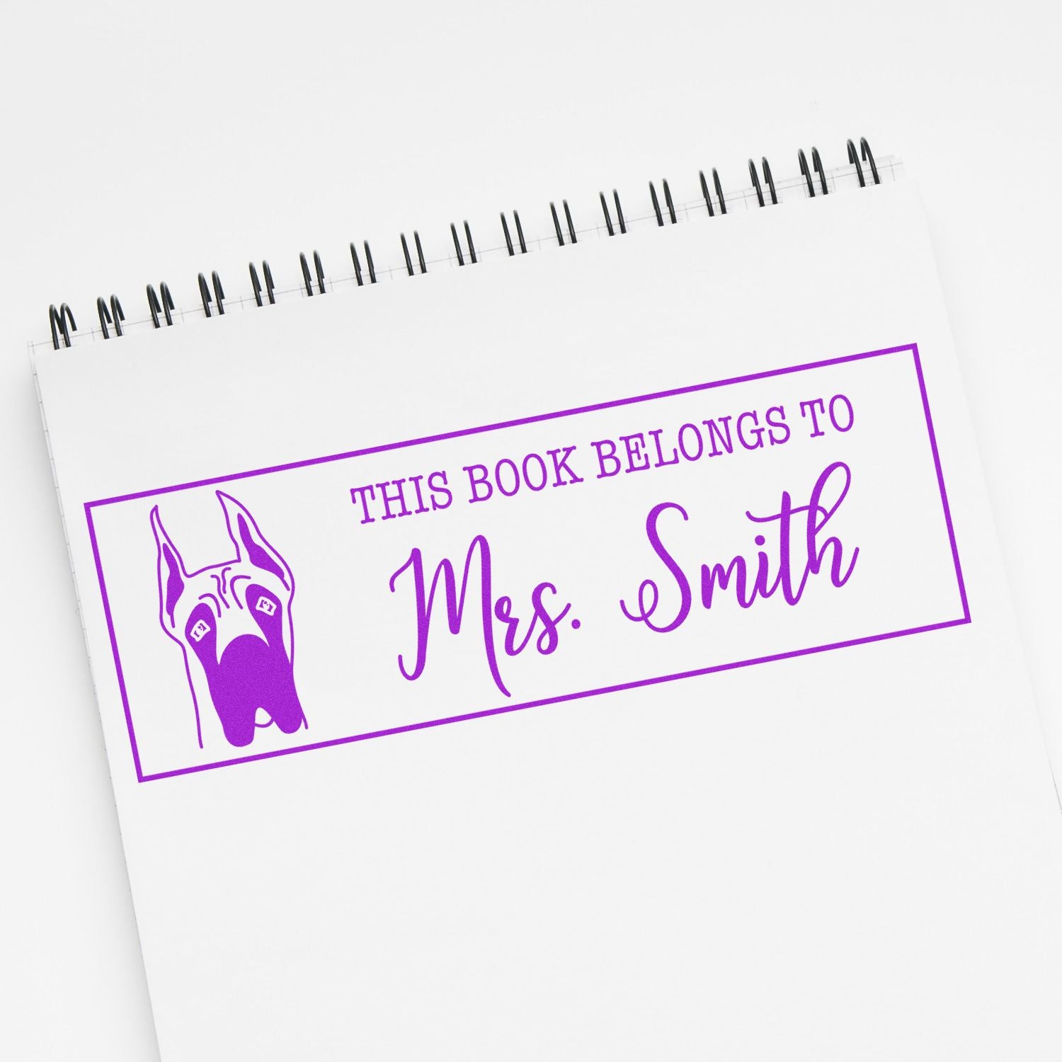 Purple custom rubber book stamp on a spiral notebook, reading This Book Belongs to Mrs. Smith with a dog illustration.