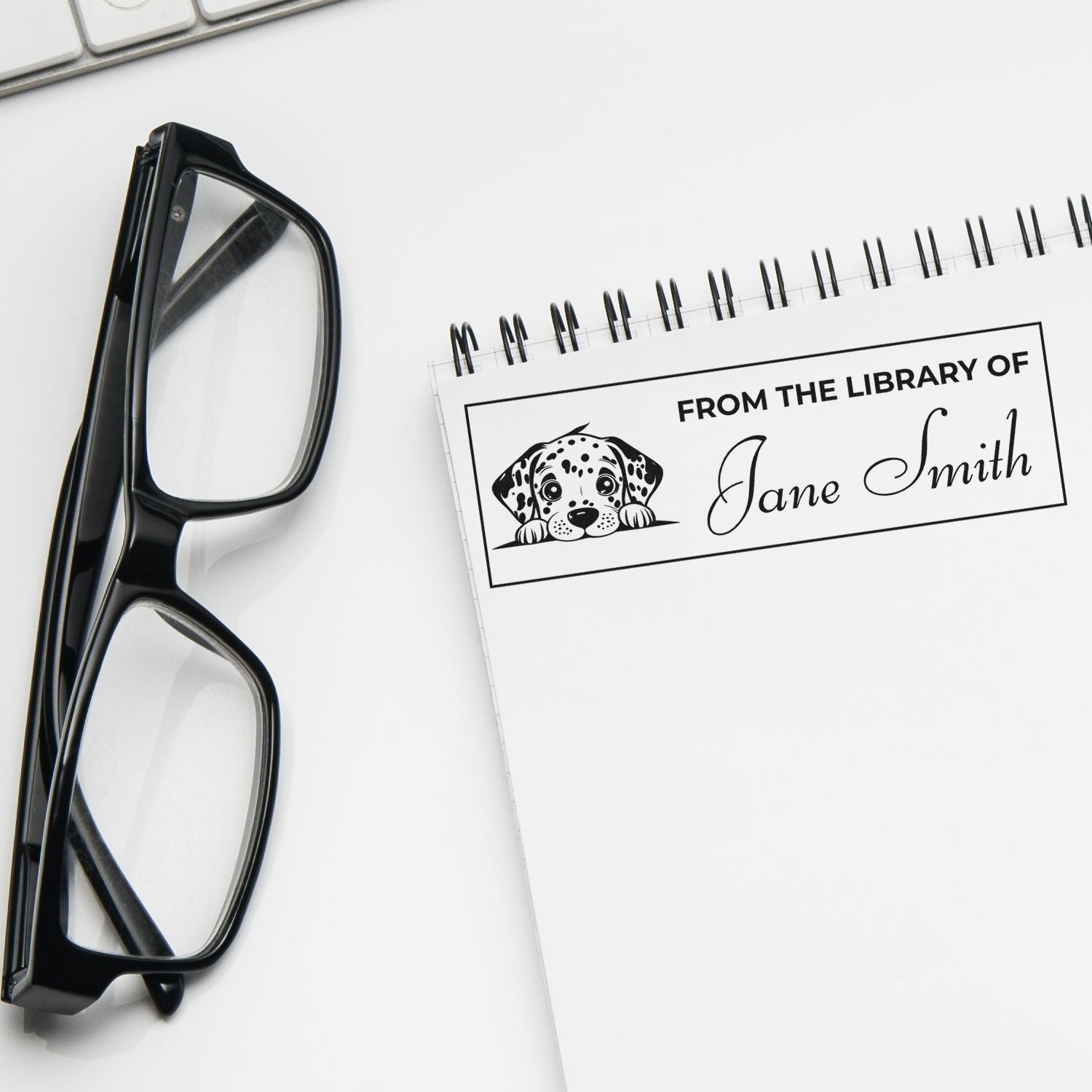 Personalized library rubber stamp with dog graphic and name, shown on a notebook beside eyeglasses for book lovers.