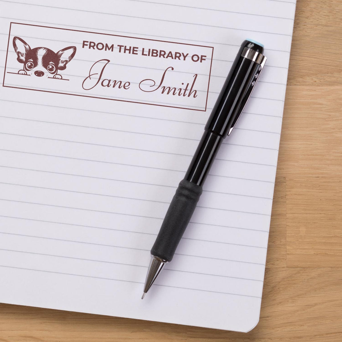Personalized library rubber stamp with dog design and From the Library of Jane Smith on a notebook beside a pen.