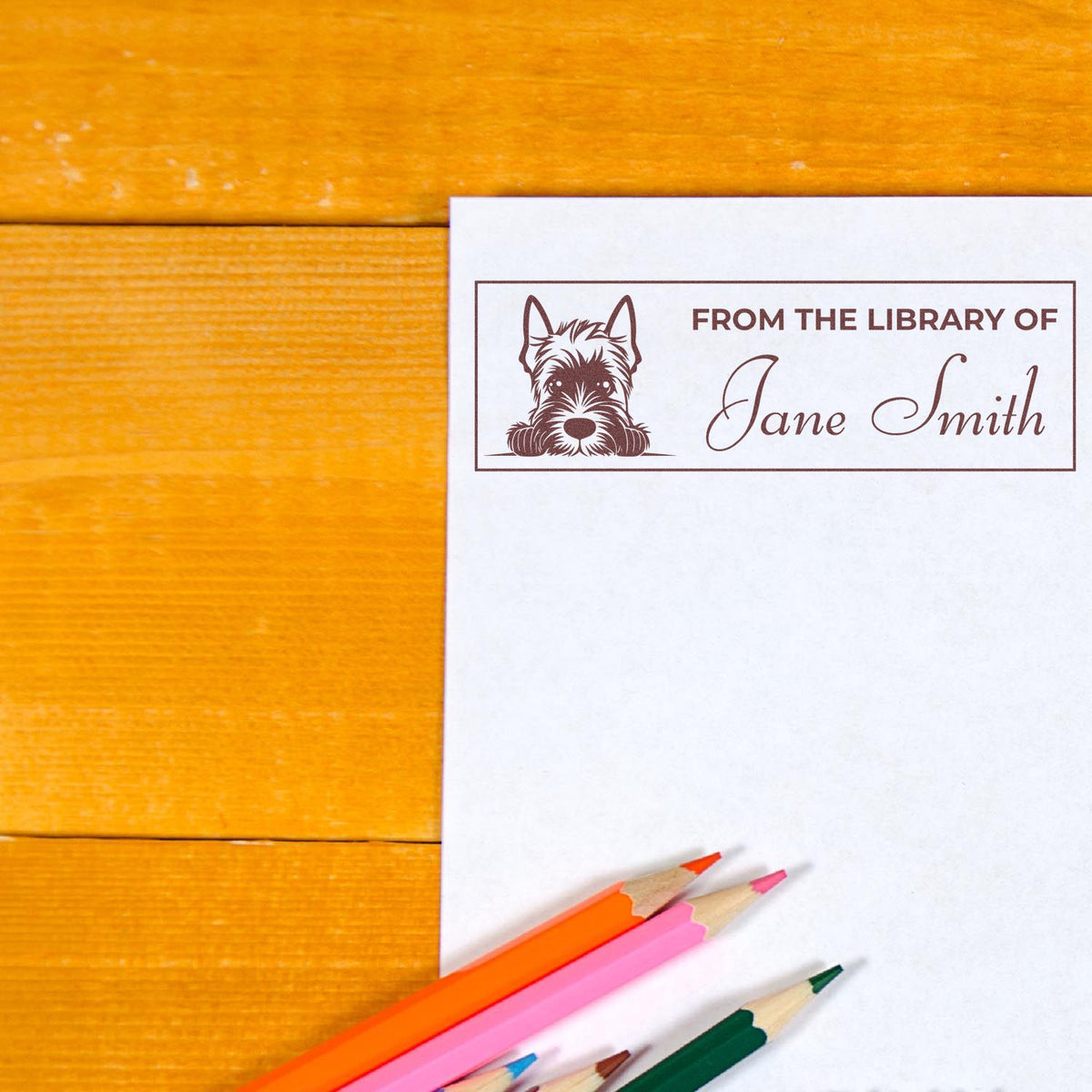 Personalized dog bookplate rubber stamp reading From the Library of Jane Smith on paper with colored pencils on a wood desk.