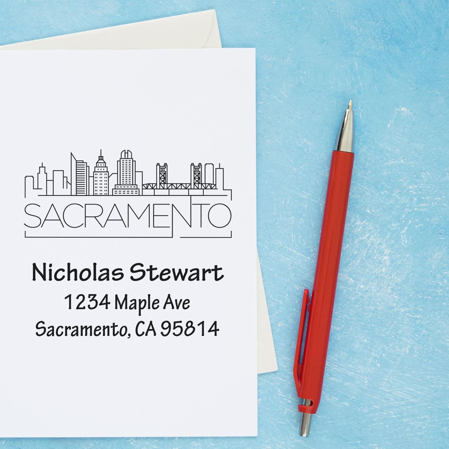 Personalized Sacramento address rubber stamp with skyline design, name, and home address on a greeting card beside a red pen.