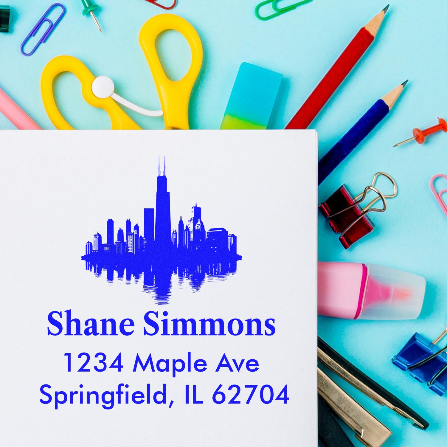 Custom skyline return address rubber stamp with name and address, displayed on white card with colorful office supplies.
