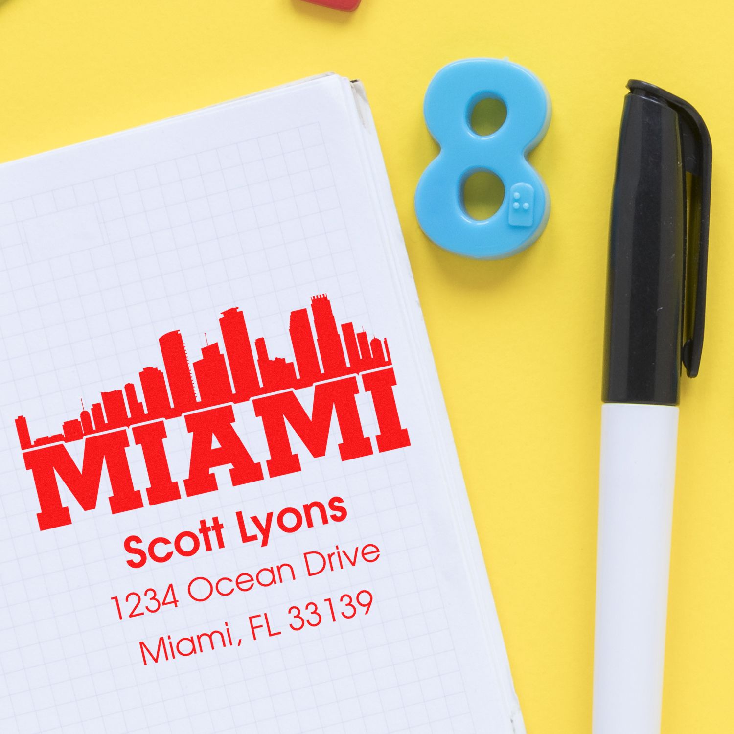 Custom Miami address rubber stamp impression on notebook, shown with pen and number 8 on yellow background.