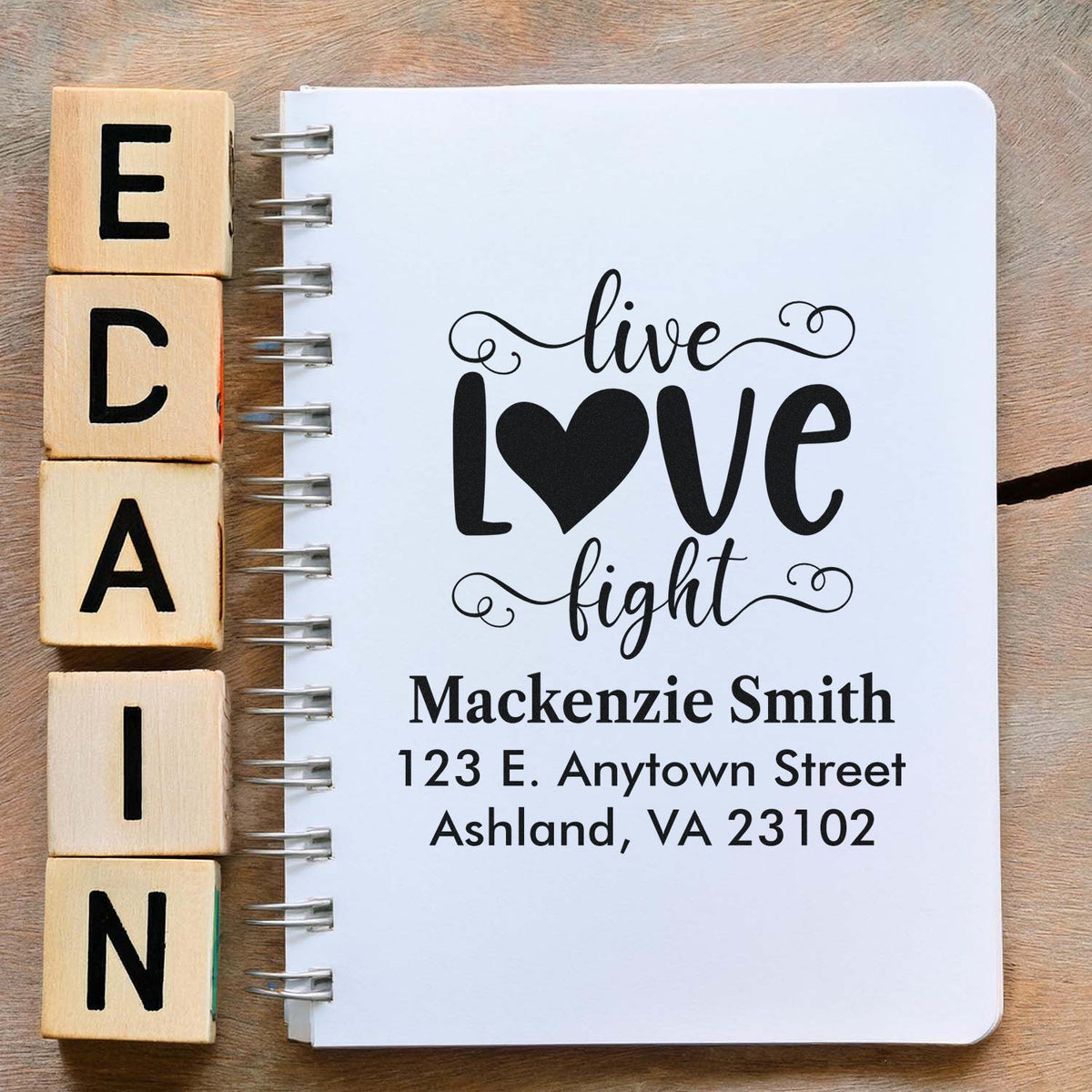 Wooden alphabet rubber stamps beside a notebook featuring a custom address stamp design with live love fight and personalized address.