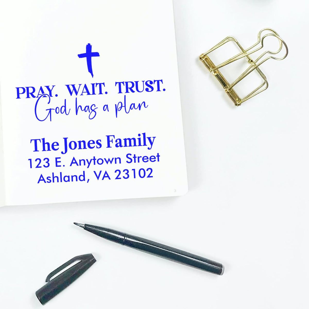 Custom rubber stamp with inspirational faith message and family return address in blue ink, shown on notebook with pen and binder clip.