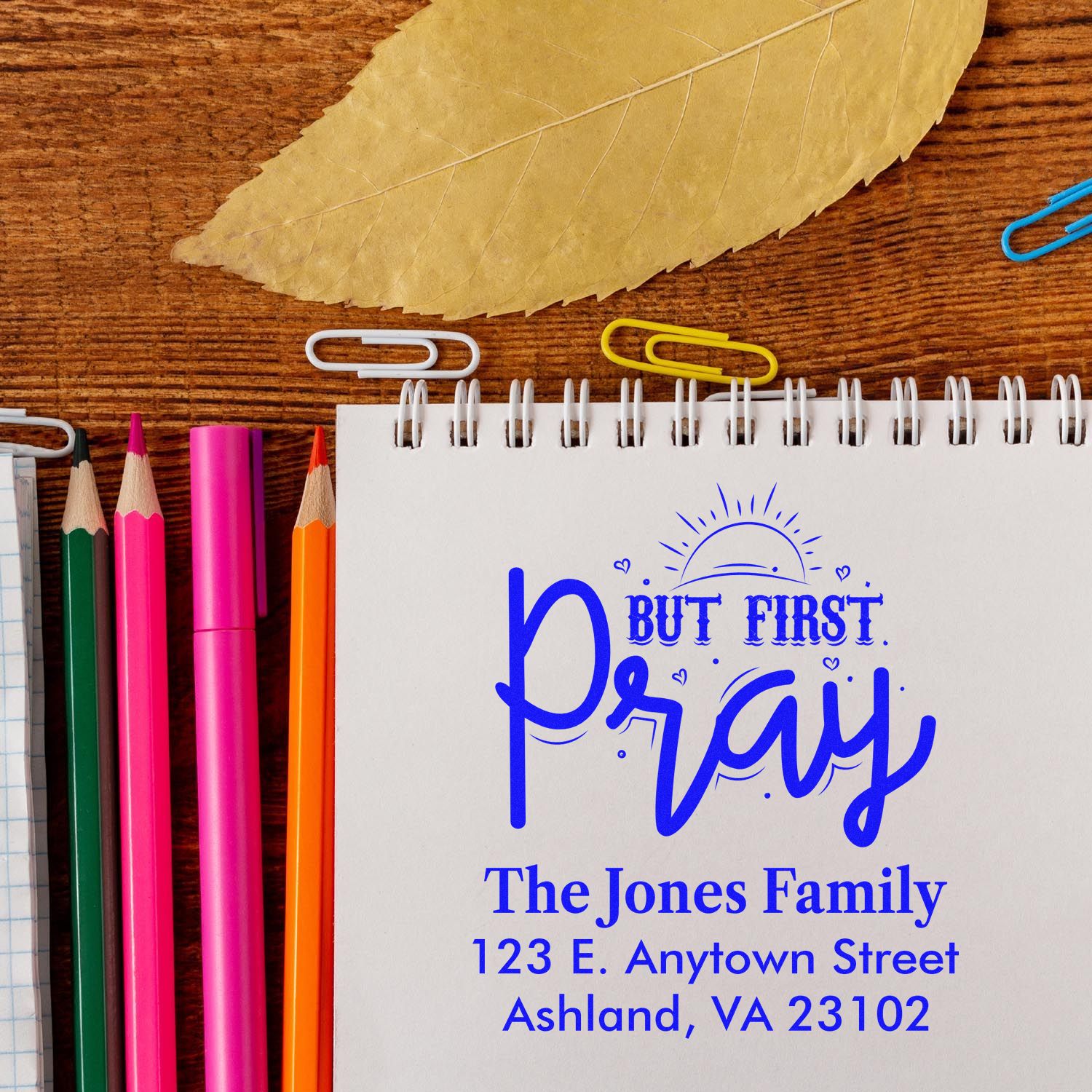 Custom rubber stamp mockup with But First, Pray return address design on notebook, with pencils and paper clips.