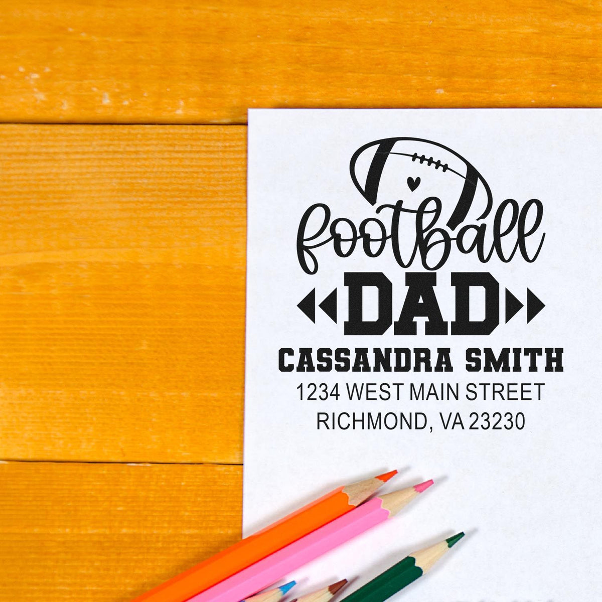 Custom football dad return address rubber stamp with name and address on paper beside colored pencils.