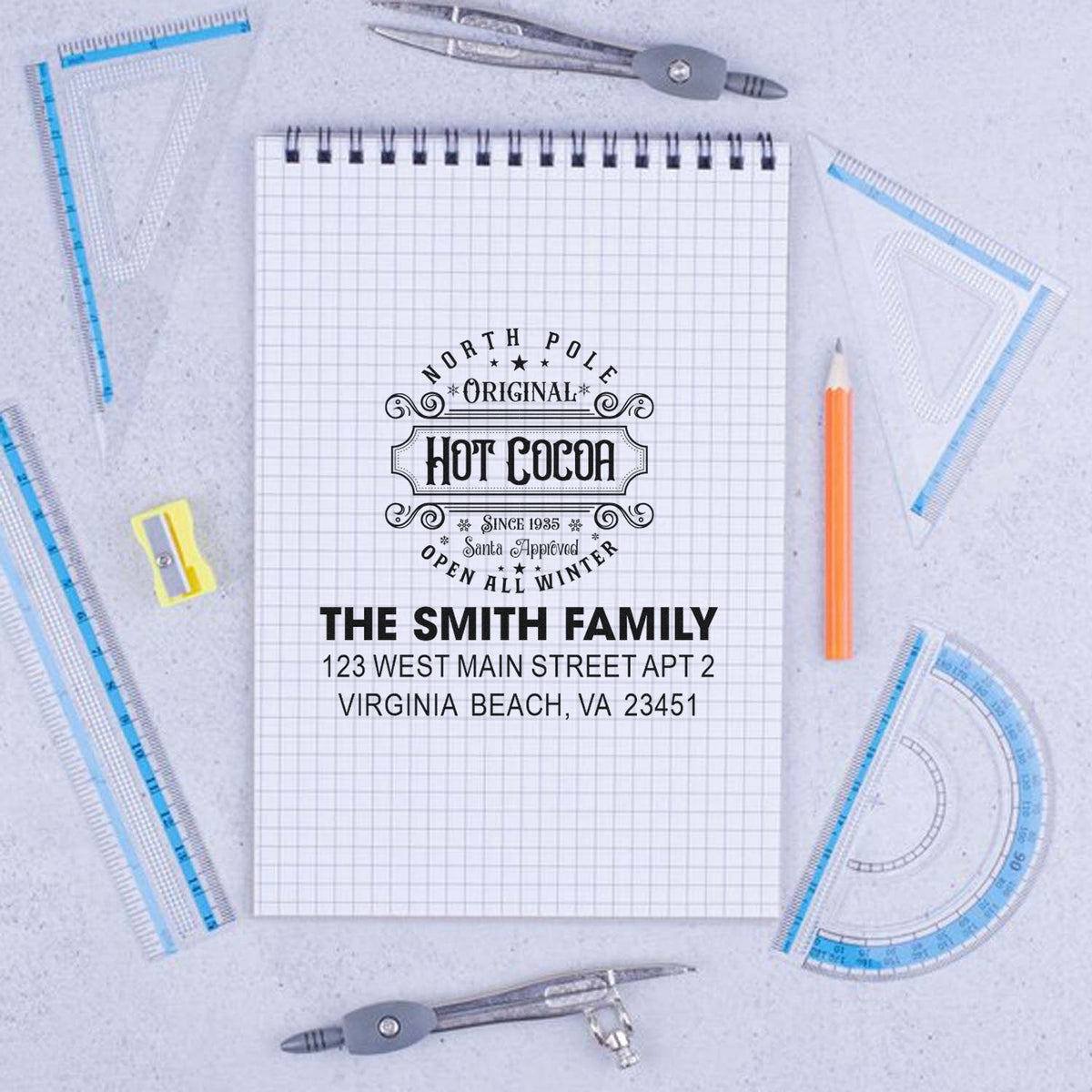 Custom rubber stamp mockup with Hot Cocoa design and Smith family address on notebook surrounded by drafting tools.