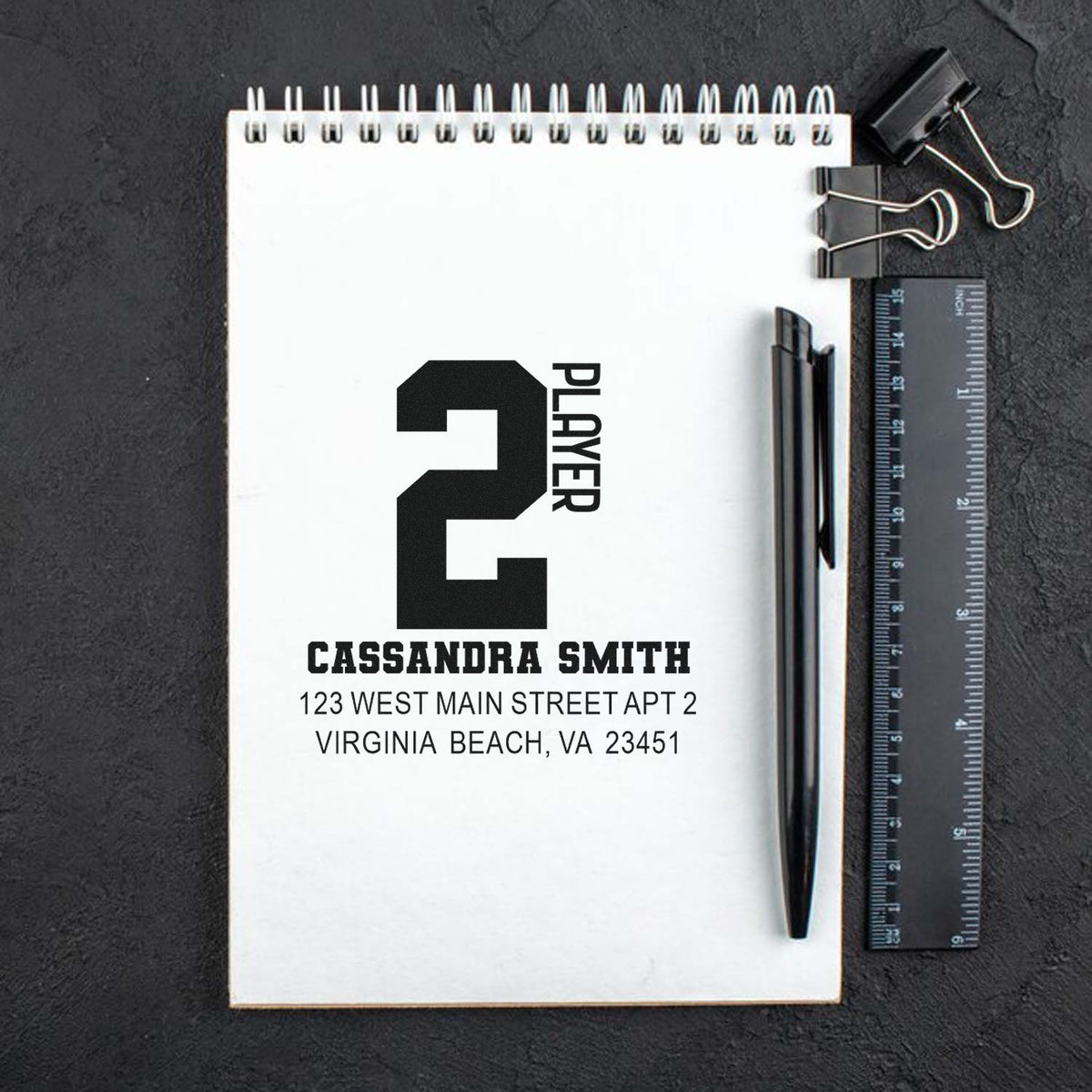 Custom rubber stamp mockup with PLAYER 2 design, name, and return address on a notebook page beside a pen and ruler.