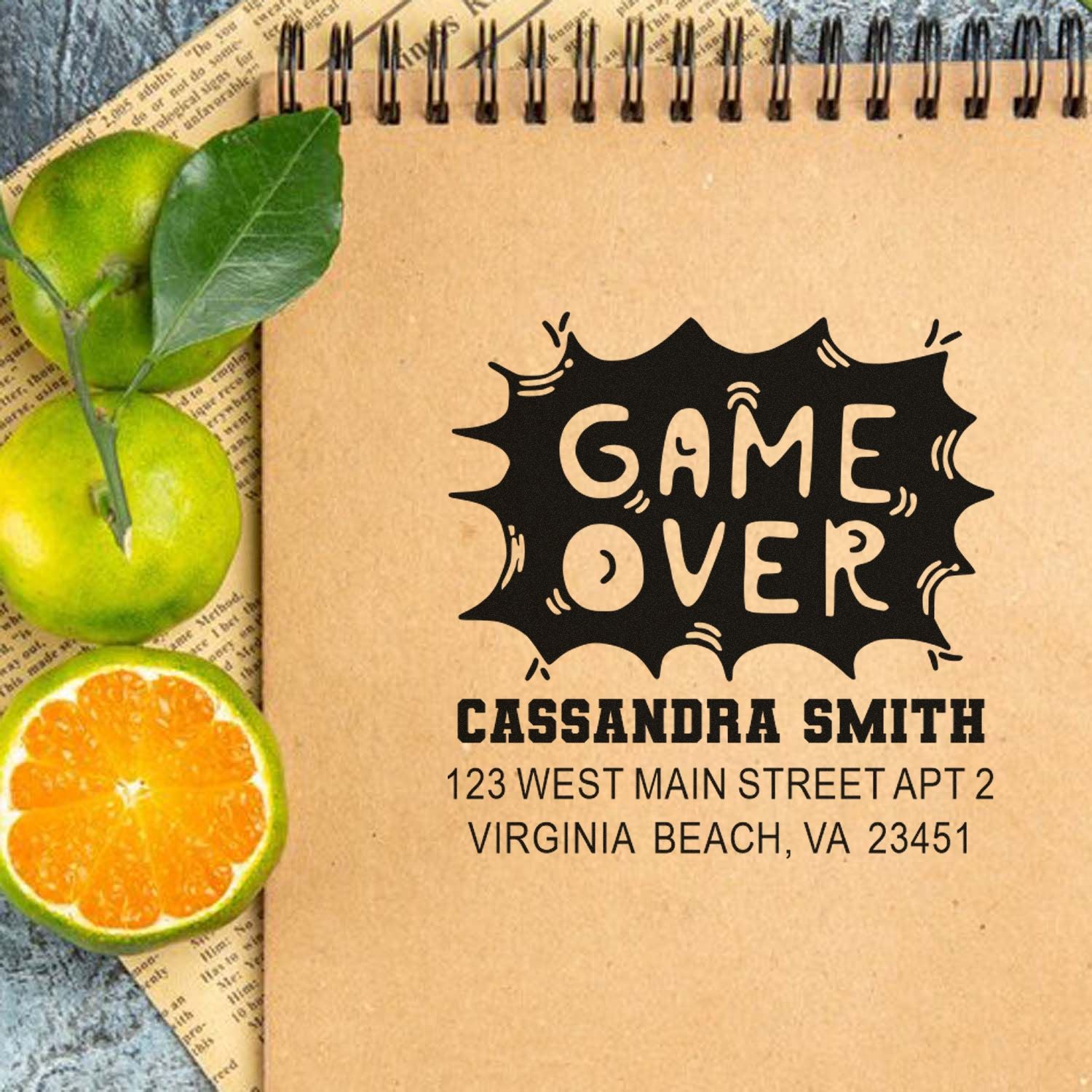 Custom Game Over address rubber stamp with personalized name and address on a kraft notebook mockup.