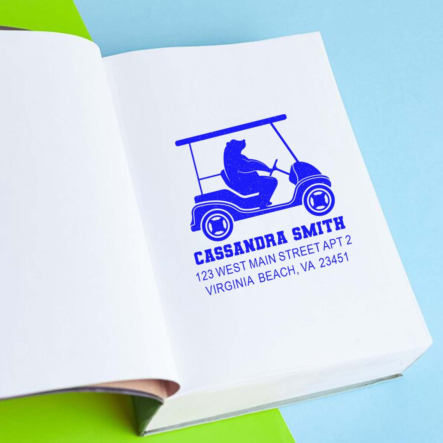 Custom rubber address stamp with golf cart logo, shown stamped in blue on a notebook page for personalized stationery branding.