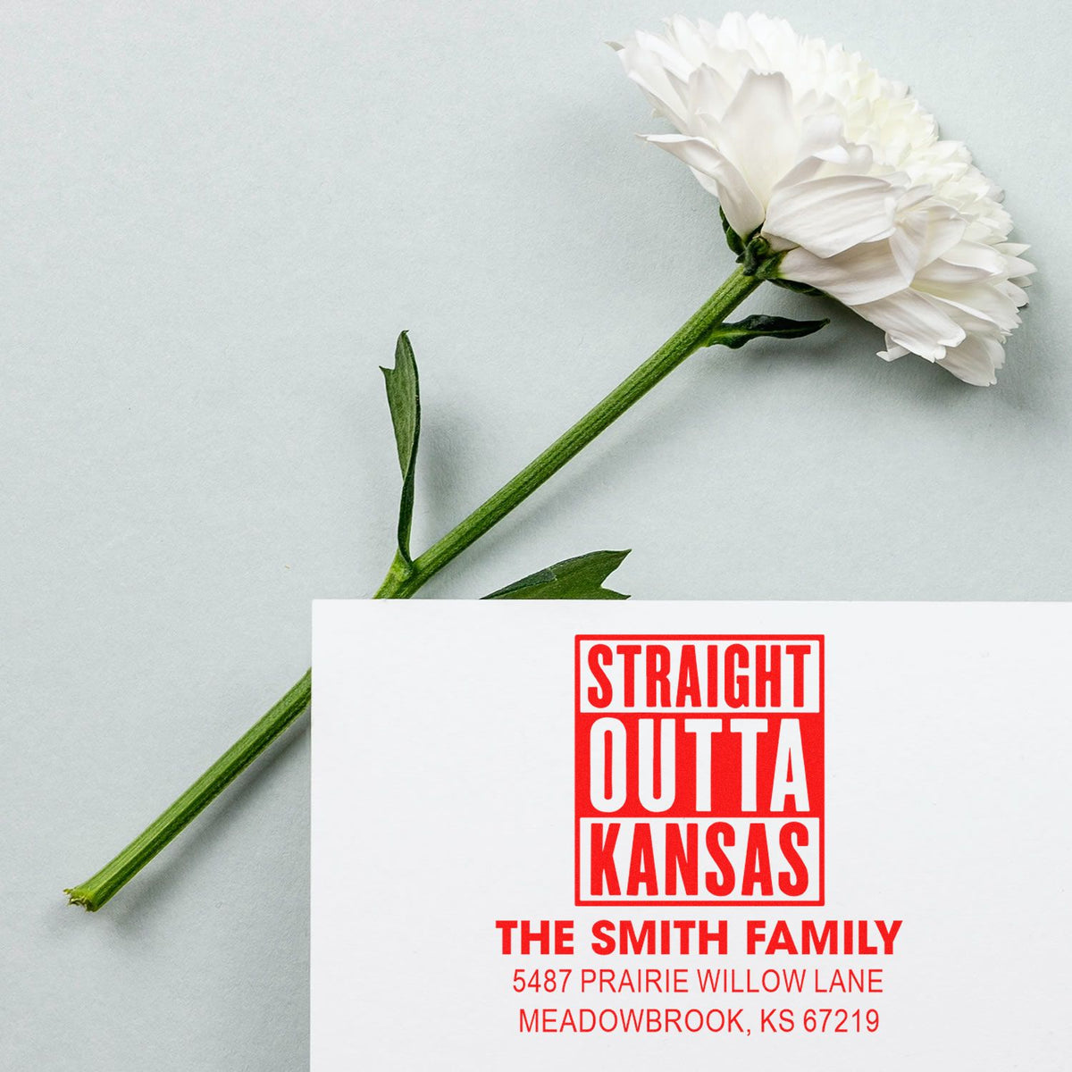 Custom address rubber stamp imprint reading Straight Outta Kansas with family name and address, shown beside a white flower.