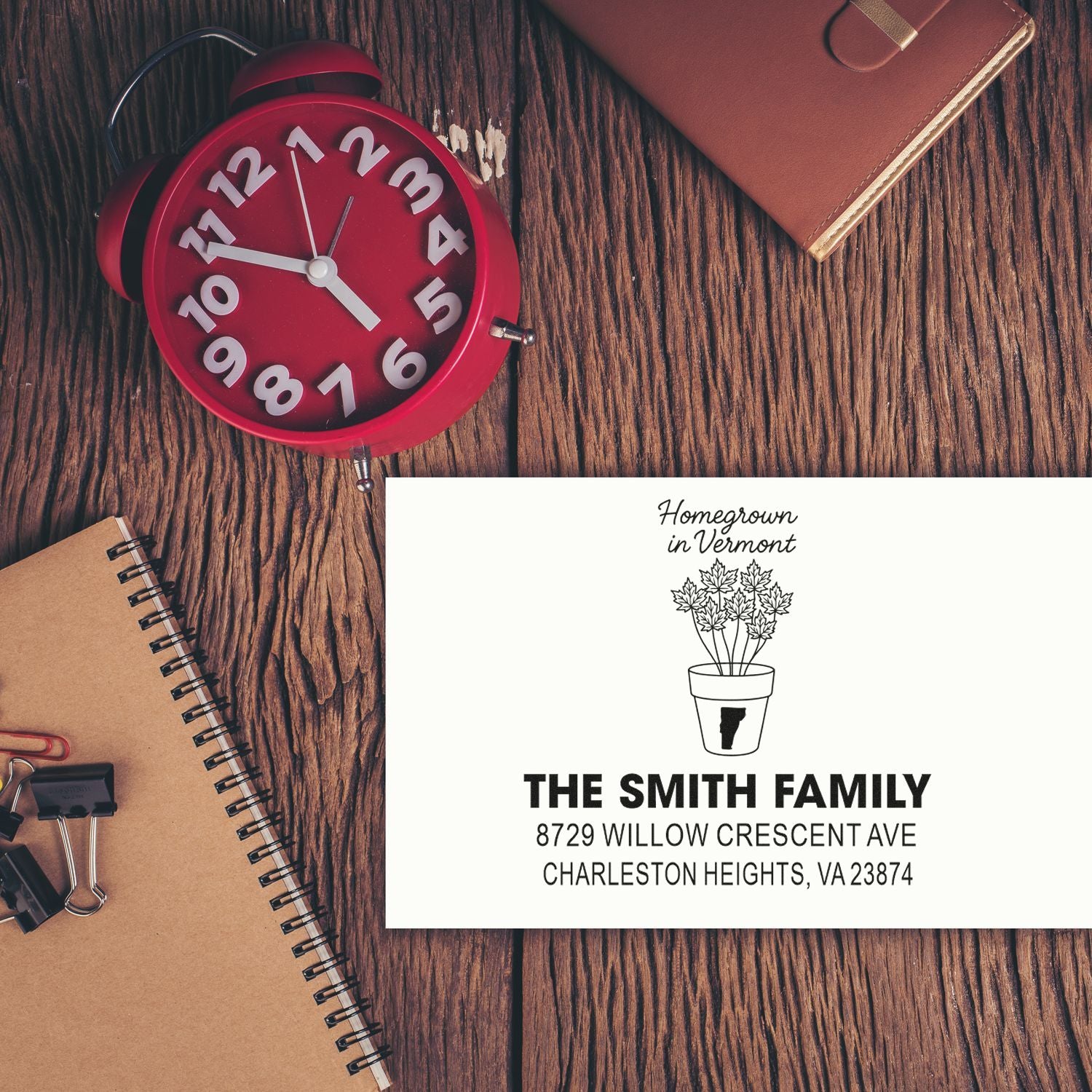 Custom self-inking address stamp with The Smith Family and potted maple design on a wooden desk beside a red alarm clock and notebook.