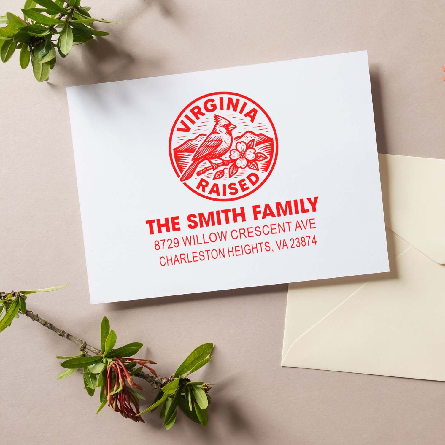 Custom red Virginia Raised family address stamp on a white card, with bird and flower design beside an envelope on a beige flat lay.