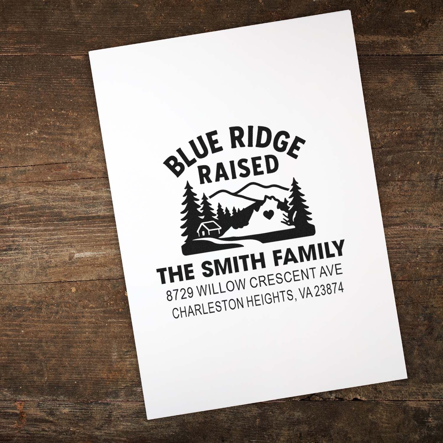 Blue Ridge Raised custom address stamp on white paper, featuring mountains, pine trees and cabin, displayed on a rustic wooden table.