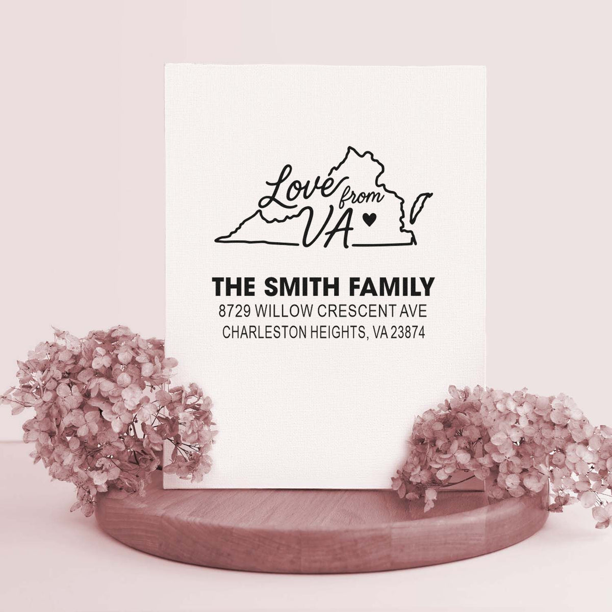 Custom Love from VA personalized address stamp for The Smith Family, displayed on a white card with pink flowers and a wooden stand.