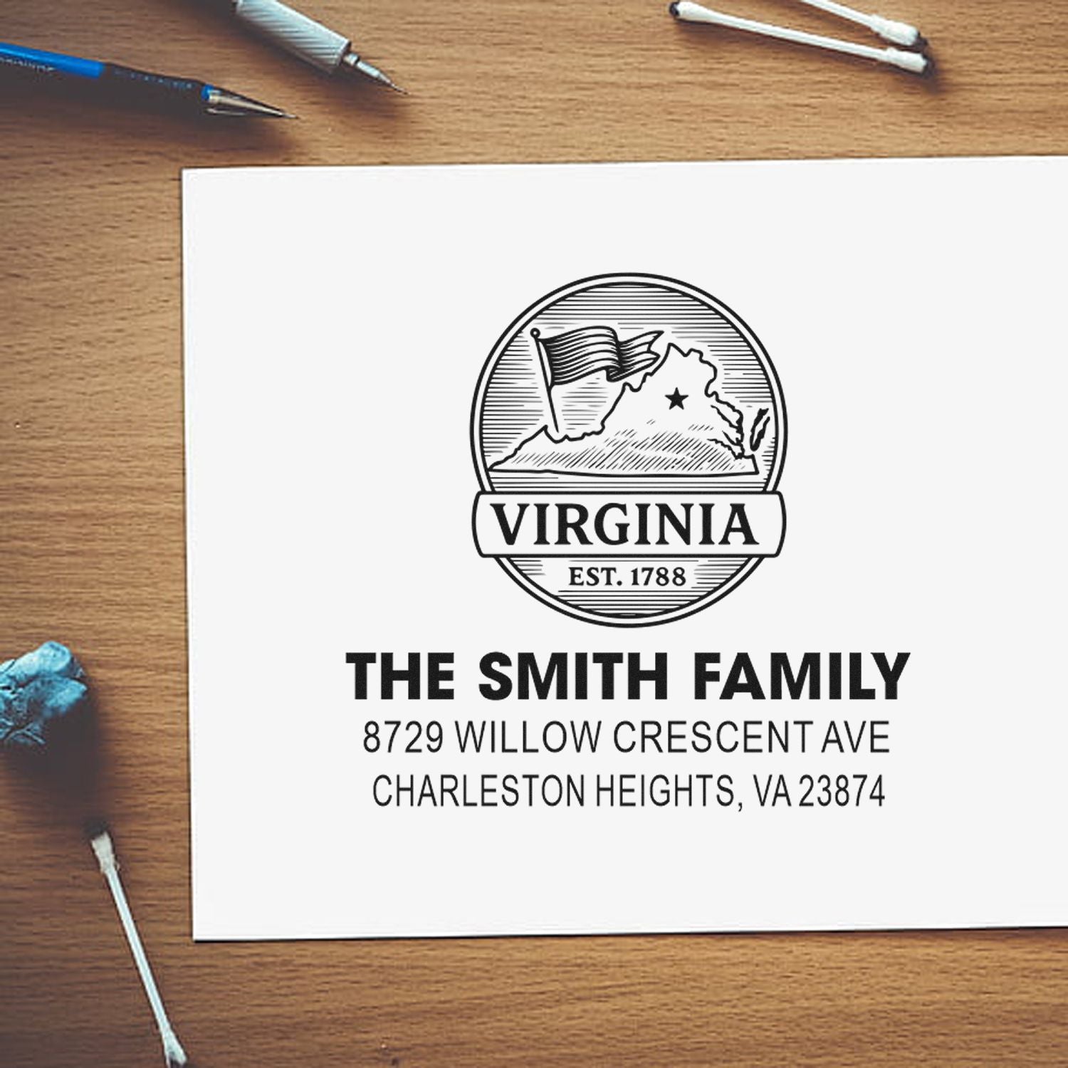 Personalized Virginia family address stamp with state emblem and home address on paper, shown on a wooden desk with pens and cotton swabs.