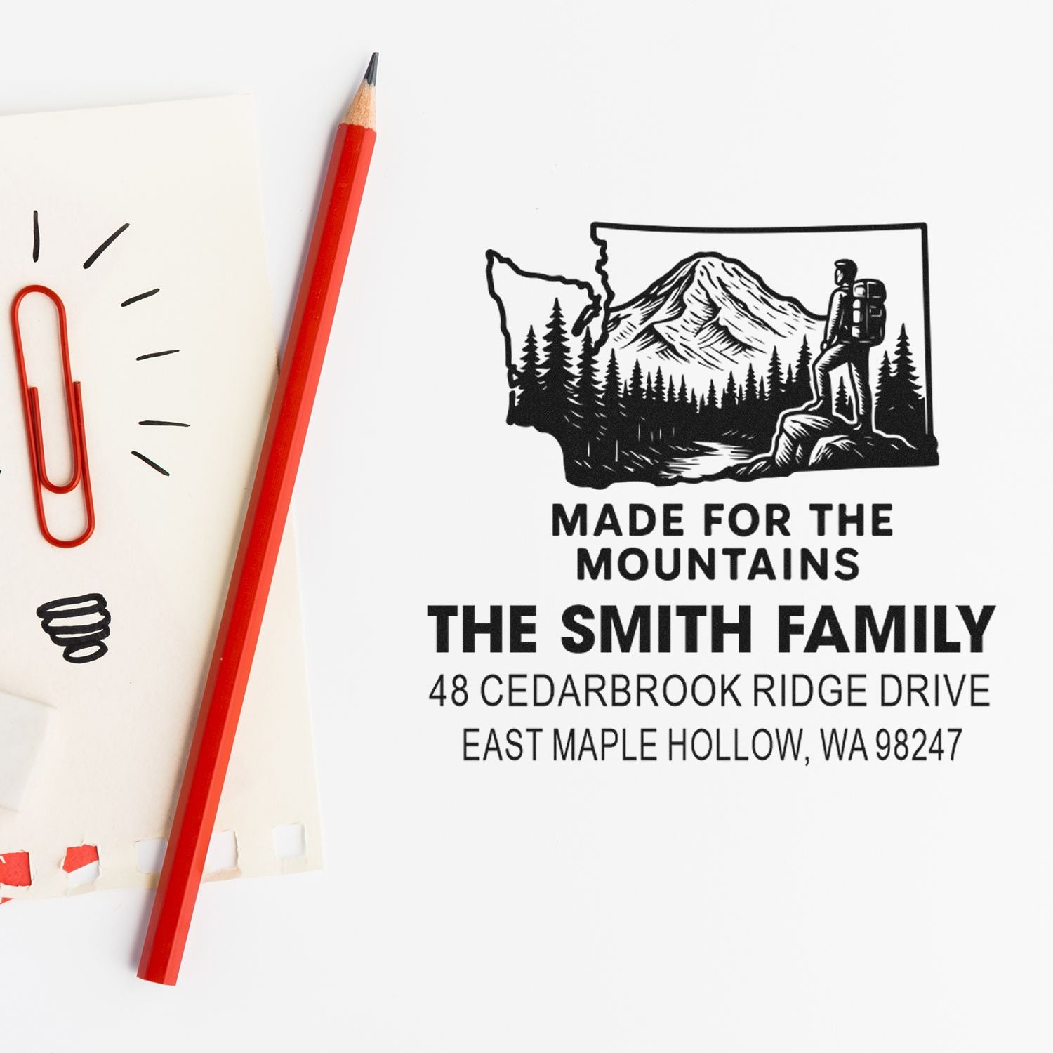 Custom address stamp with mountain hiker design, plus pencil and paper clips on a white desk, reading Made for the Mountains for The Smith Family.